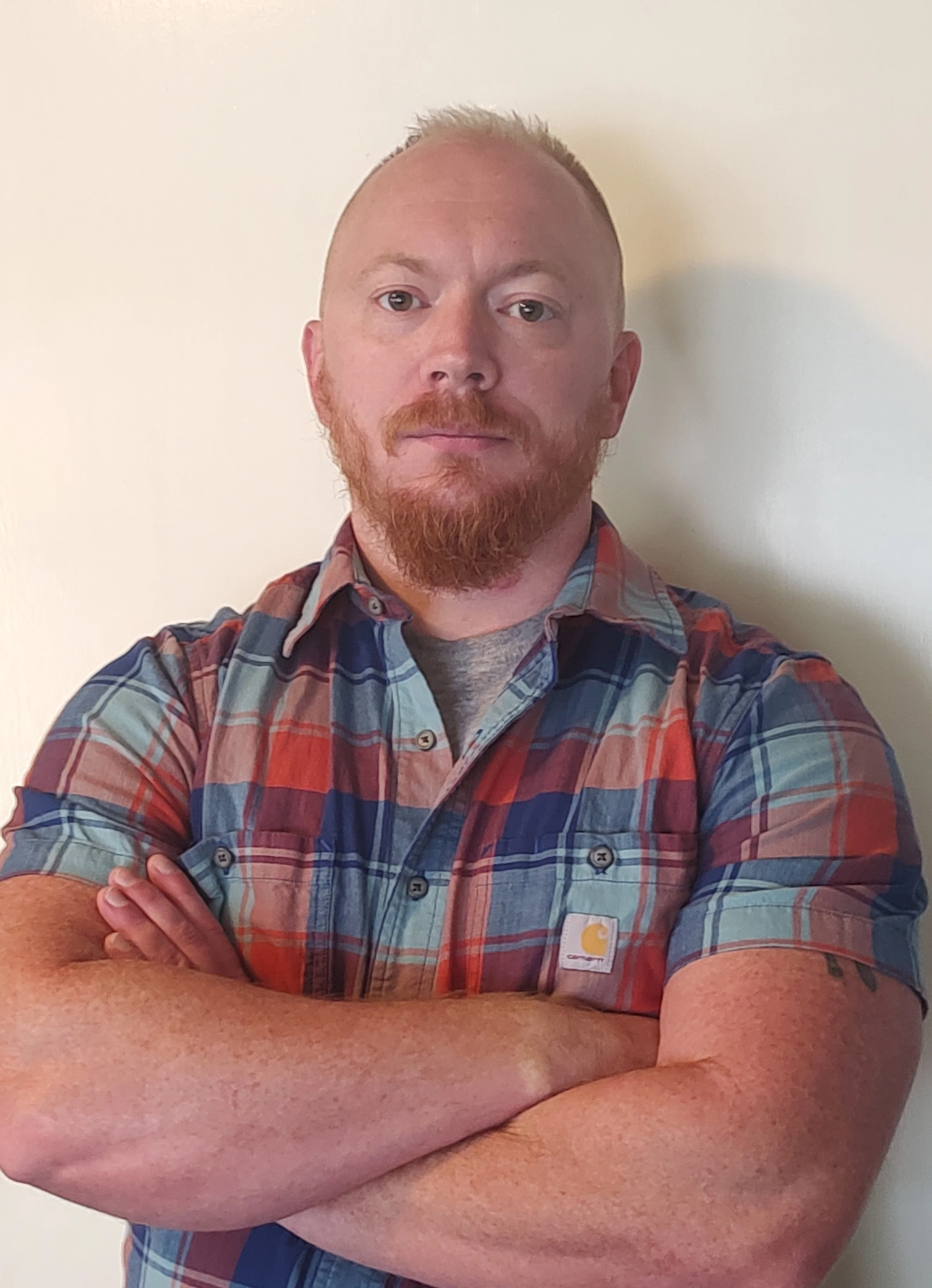 A man with red hair and beard wearing a colorful plaid shirt, arms crossed, standing against a plain light-colored wall.