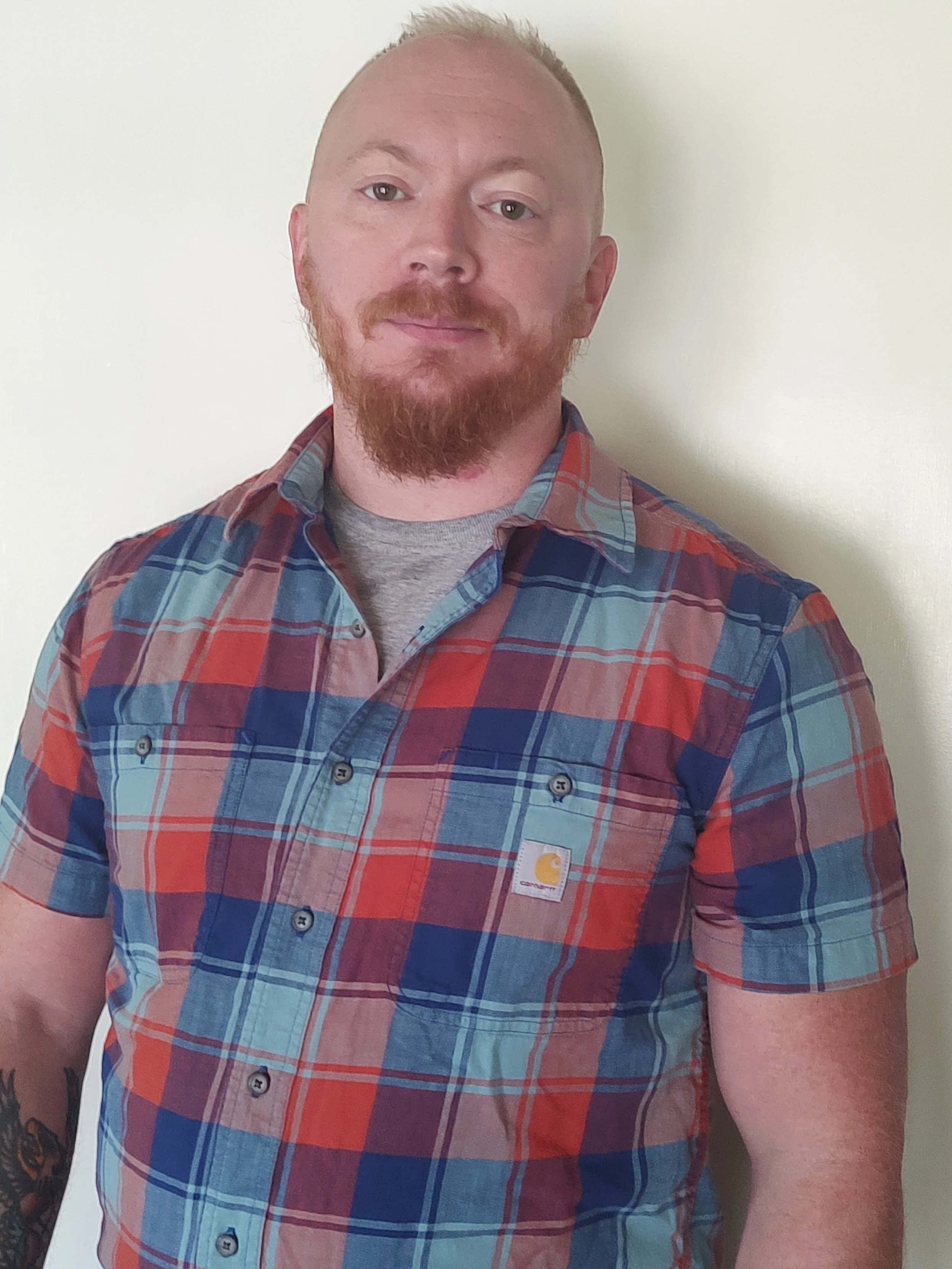 A man with short blonde hair, a reddish beard, wearing a multicolored plaid short-sleeve shirt with a Carhartt logo, standing against a plain off-white wall.