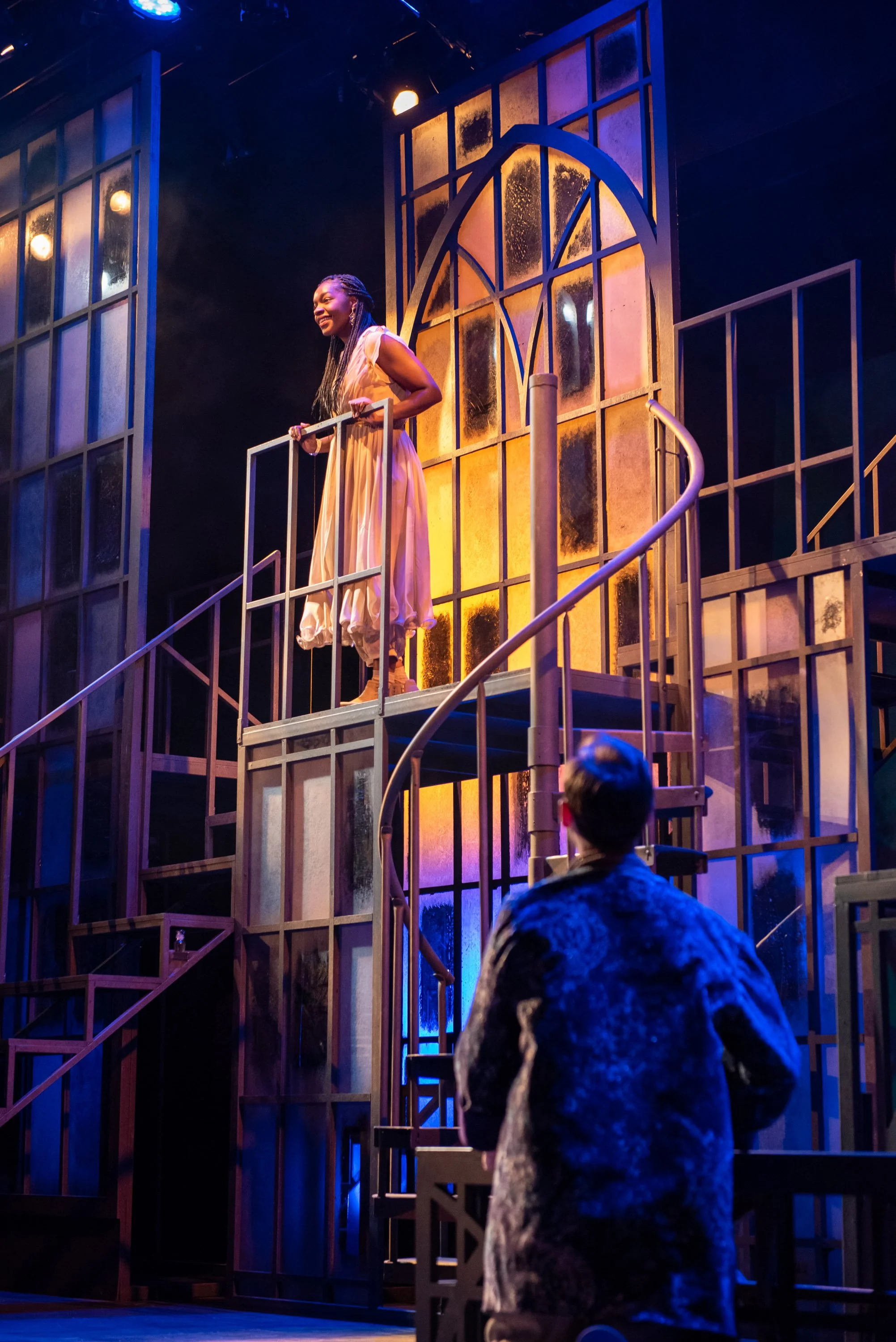 Romeo and Juliet at the Barter Theatre of Abingdon, VA.  A woman on stage, smiling and holding onto a railing, wearing a long light-colored dress, with a backdrop resembling a cathedral window structure, illuminated by stage lights.