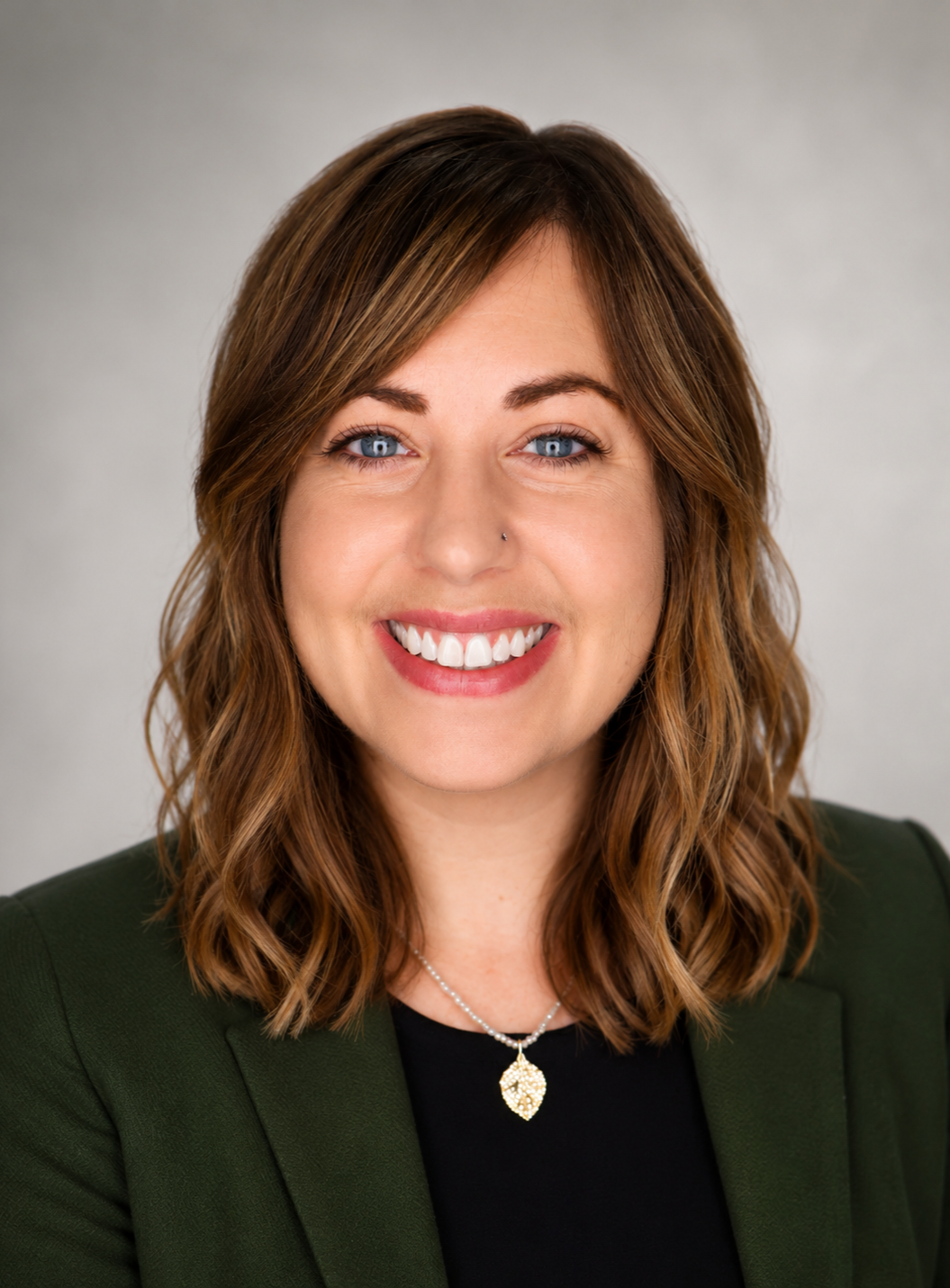 Headshot of Megan Randall with a gray background. She is white, has shoulder-length wavy brown hair, and blue eyes. She is wearing a dark green blazer, a black top, and a white beaded necklace with a gold leaf-shaped pendant.