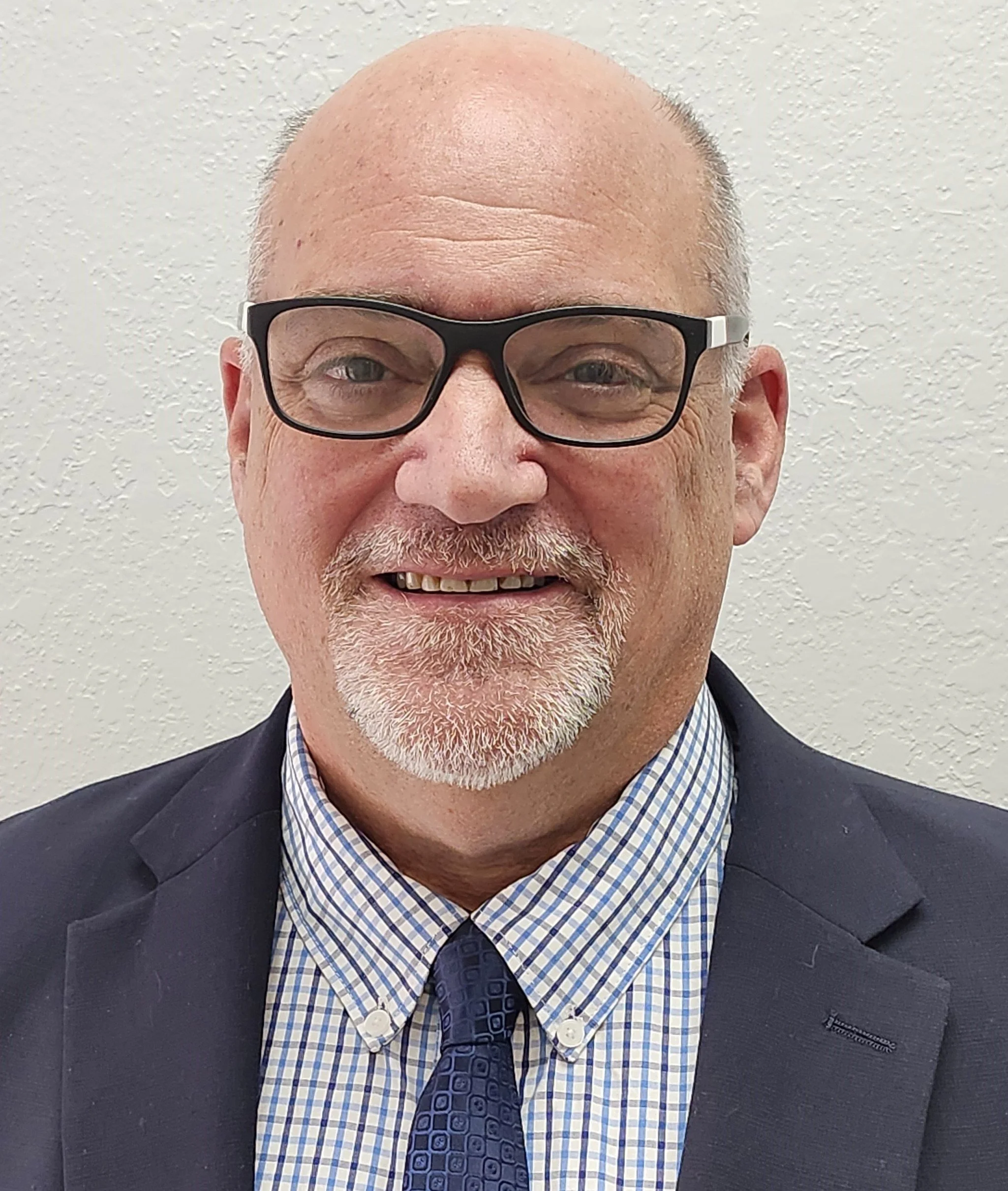Close-up of smiling middle-aged man with glasses, a checked shirt, a tie, and a dark blazer, standing against a plain textured wall.