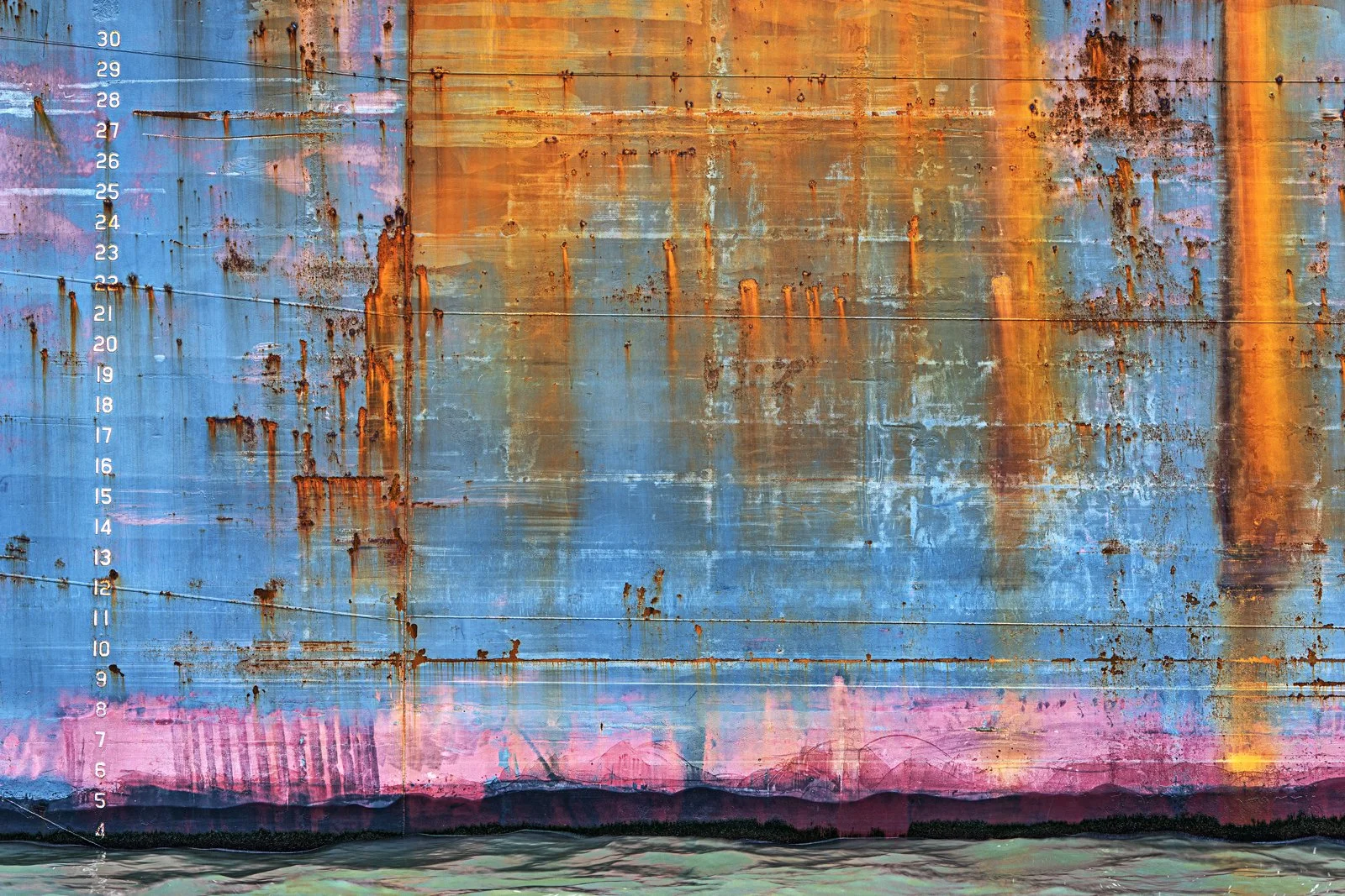 A weathered, rusted ship hull painted in a vibrant swirl of colors, including blue, orange, pink, and purple, with a waterline at the bottom and a height measurement scale on the left side.
