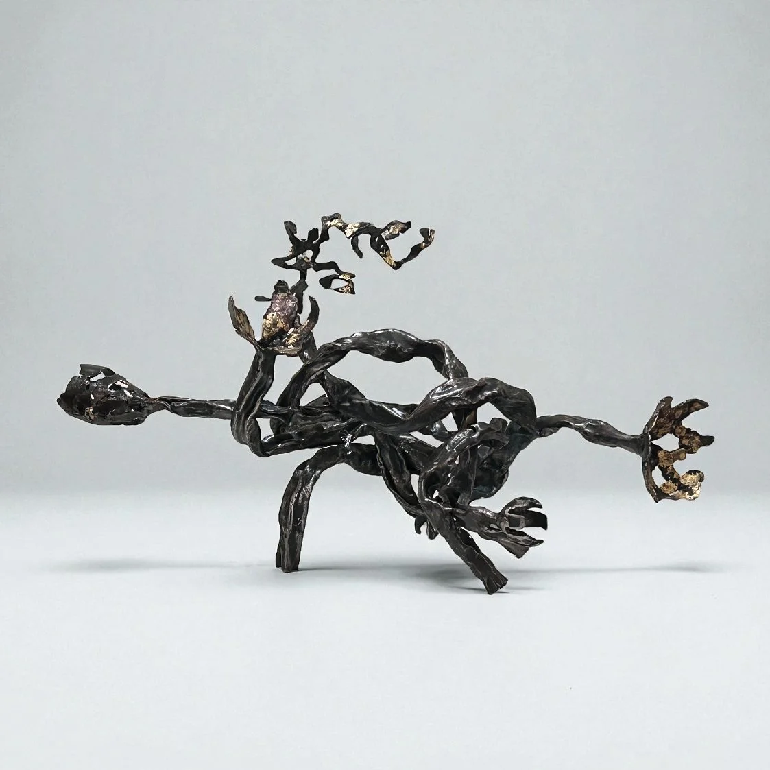 Metal sculpture of a dog in a running pose, made of dark, twisted metal with gold accents, set against a plain, light background.