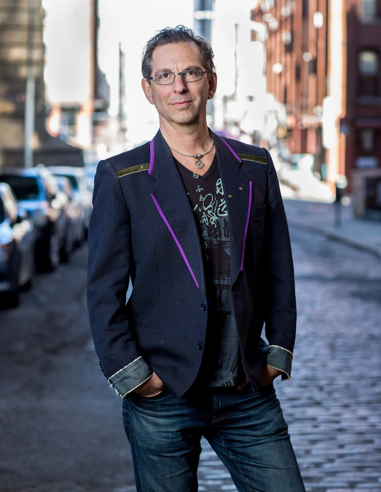 Man standing on cobblestone street in city, wearing glasses, a black t-shirt with white designs, a black blazer with purple accents, and jeans, with hands in pockets and hands in pockets, with blurred buildings and parked cars in background.