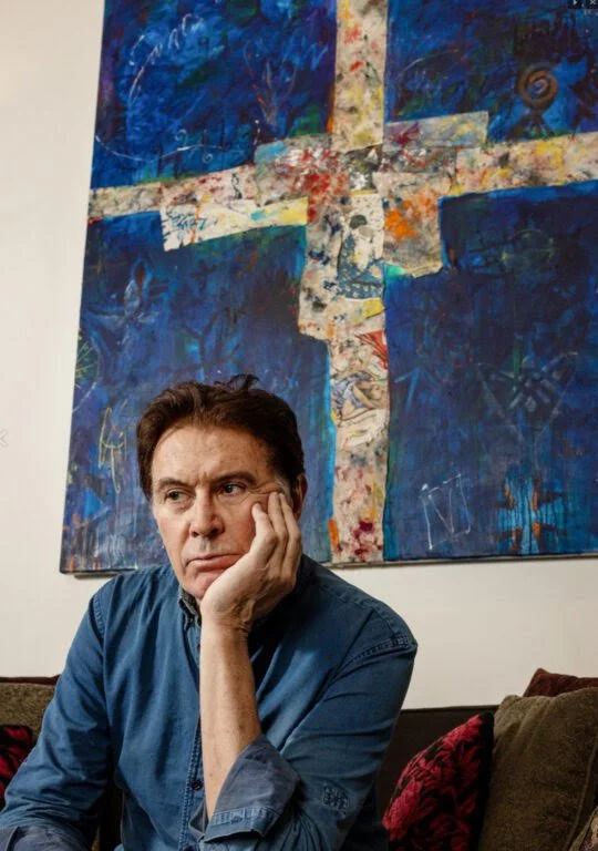 A man with dark hair and a serious expression, resting his head on his hand, sitting on a sofa with colorful pillows, in front of an abstract painting with vibrant blue, white, red, and orange colors on a white wall.
