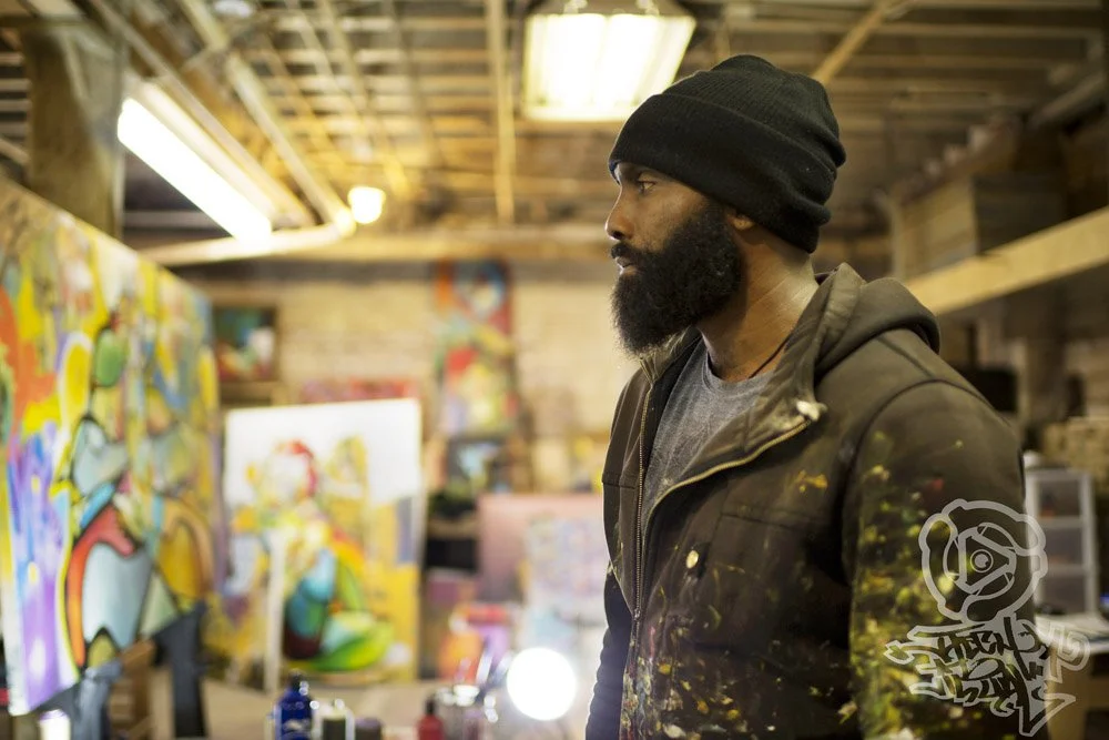 A bearded man wearing a black beanie and a dark jacket with paint splatters, standing in an art studio surrounded by colorful paintings and art supplies.