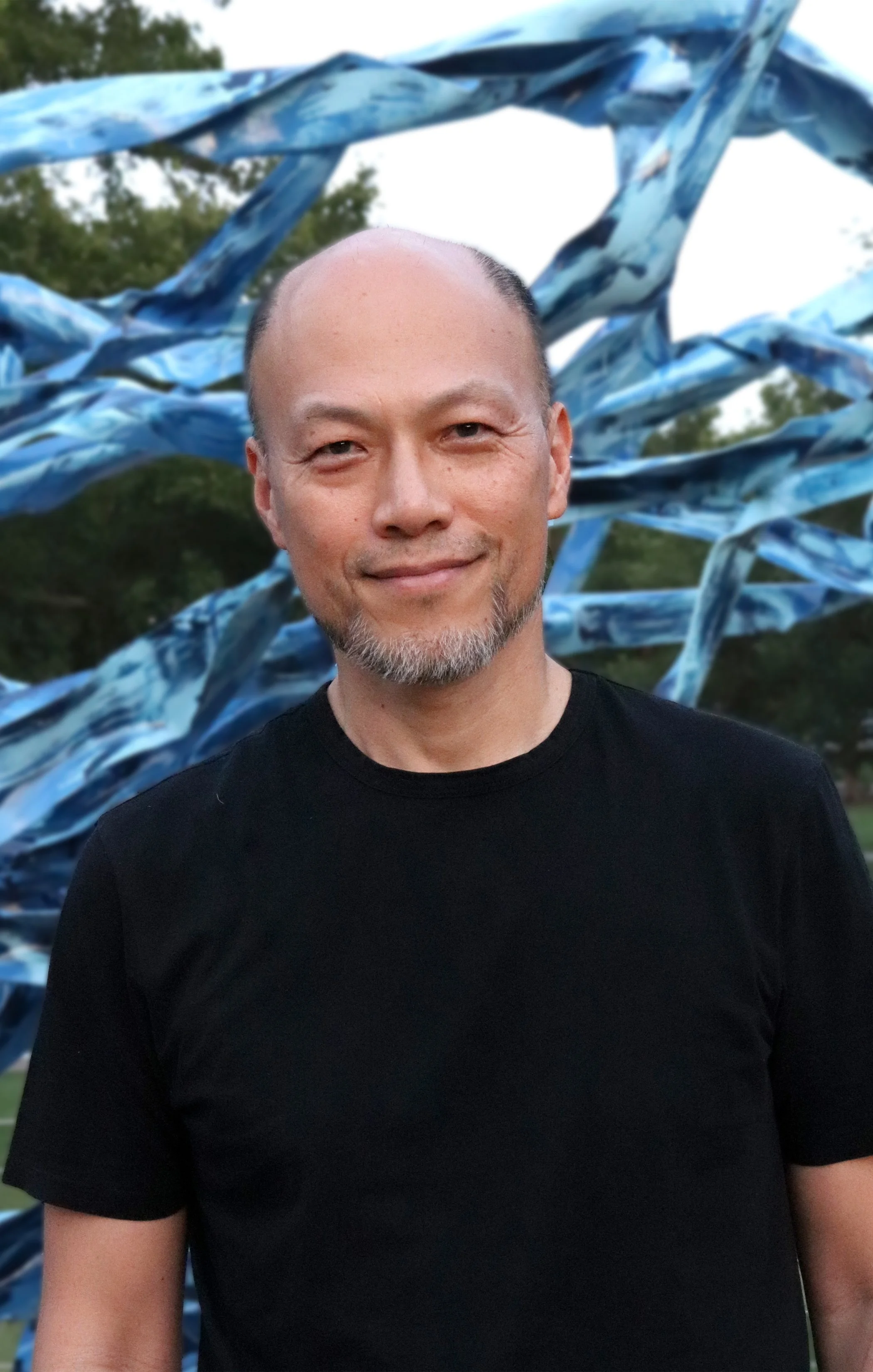 A man with a goatee smiling in front of a blue and white abstract sculpture outdoors.