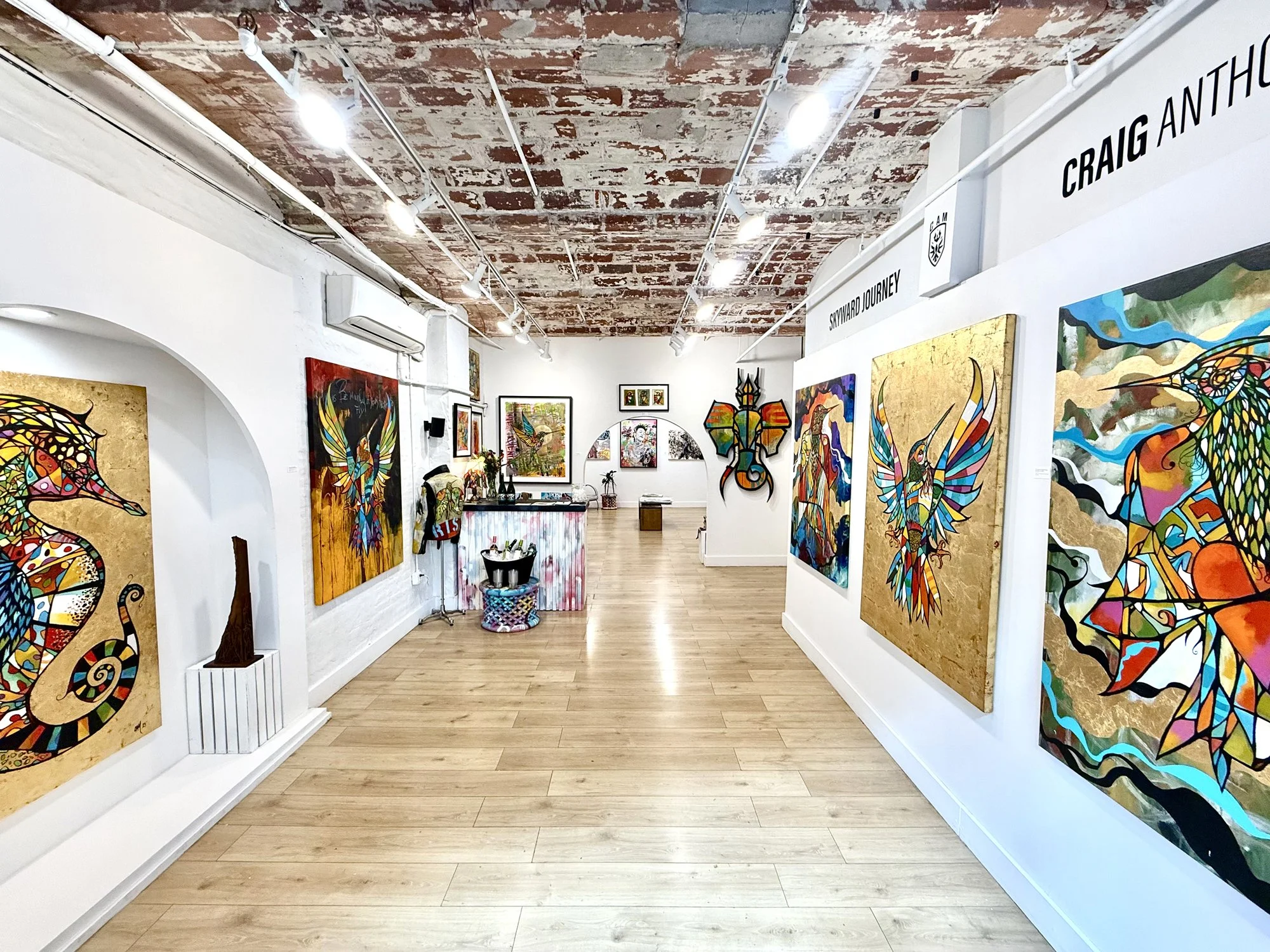 Art gallery with colorful abstract paintings of dragons and other designs on white walls, wooden floor, exposed brick ceiling, and track lighting.