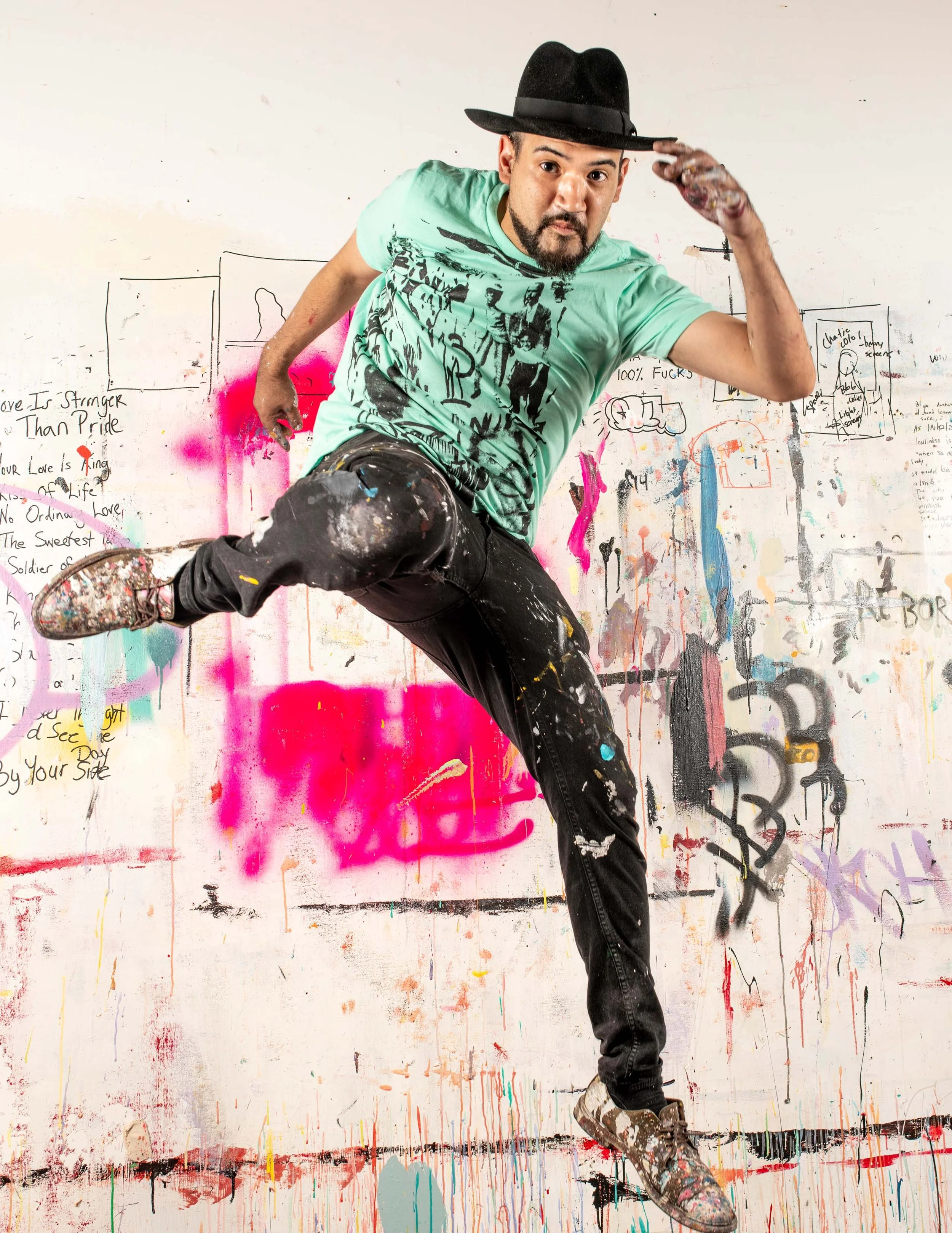 A man in a black hat and casual clothes poses dynamically in front of a colorful, graffiti-covered wall.
