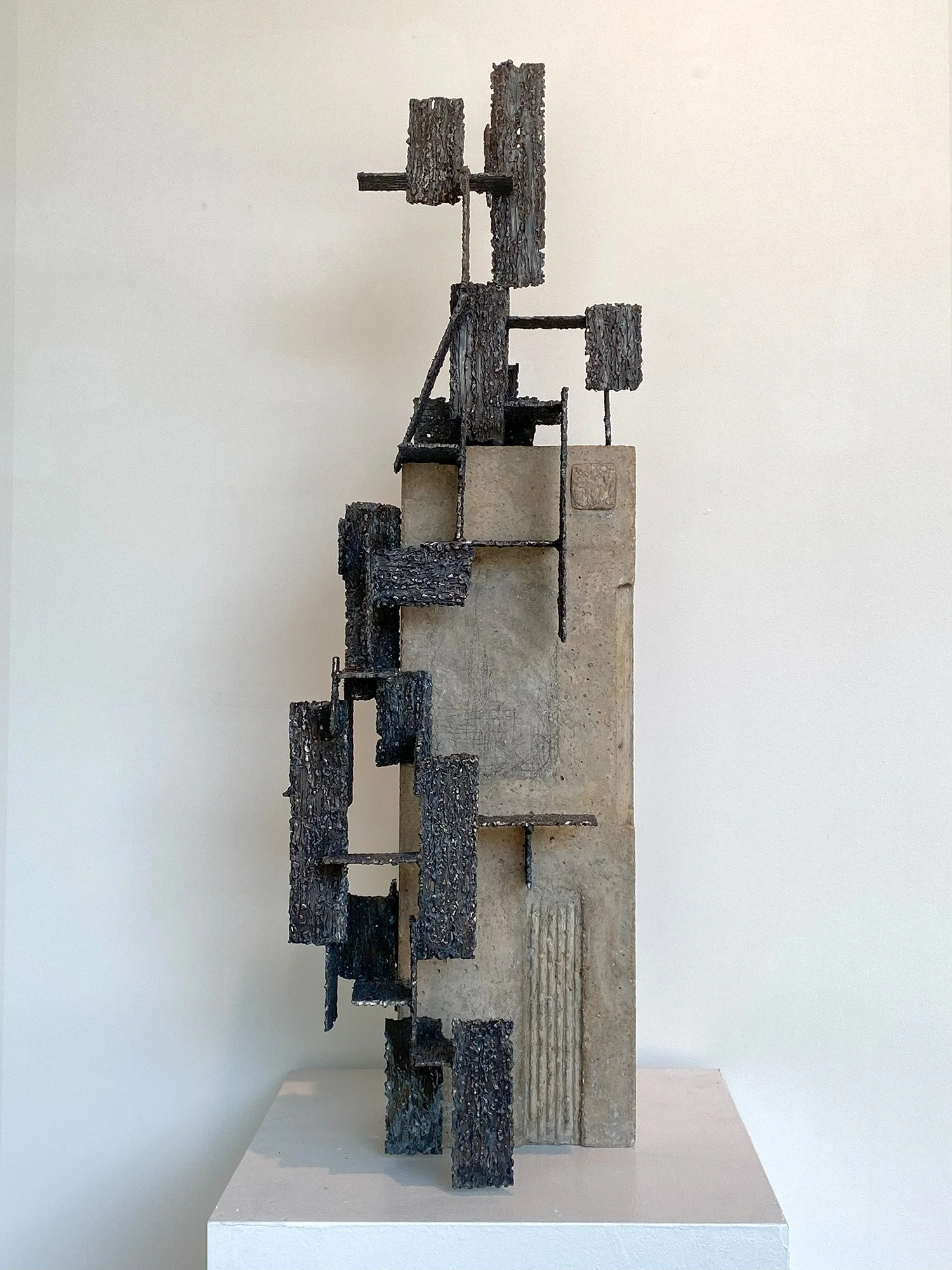 A modern sculpture composed of vertically oriented blackened wood pieces and metal rods, mounted on a white pedestal against a white wall background.