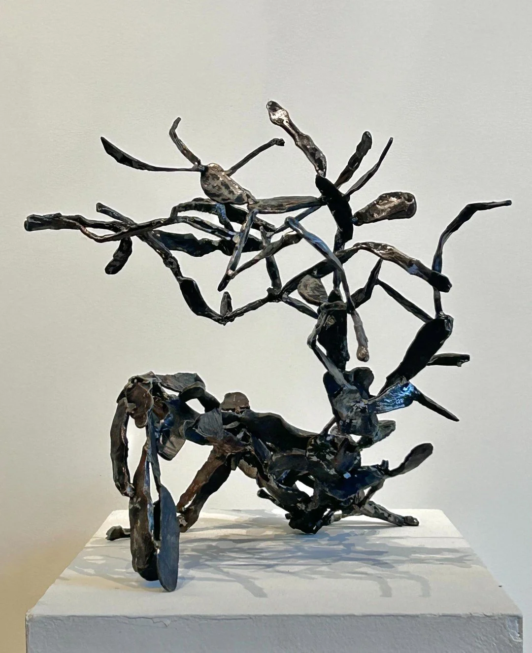 A metal sculpture of a dragonfly perched on a branch, displayed on a white pedestal against a plain background.