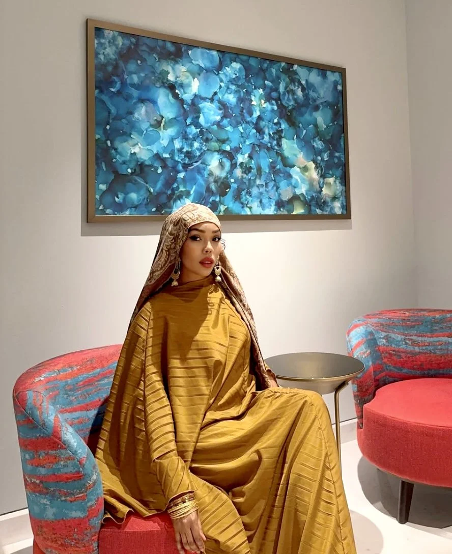 A woman wearing traditional or cultural attire in a mustard yellow dress and matching headscarf, sitting on a colorful chair in an art gallery or modern interior space, with abstract blue artwork on the wall behind her.