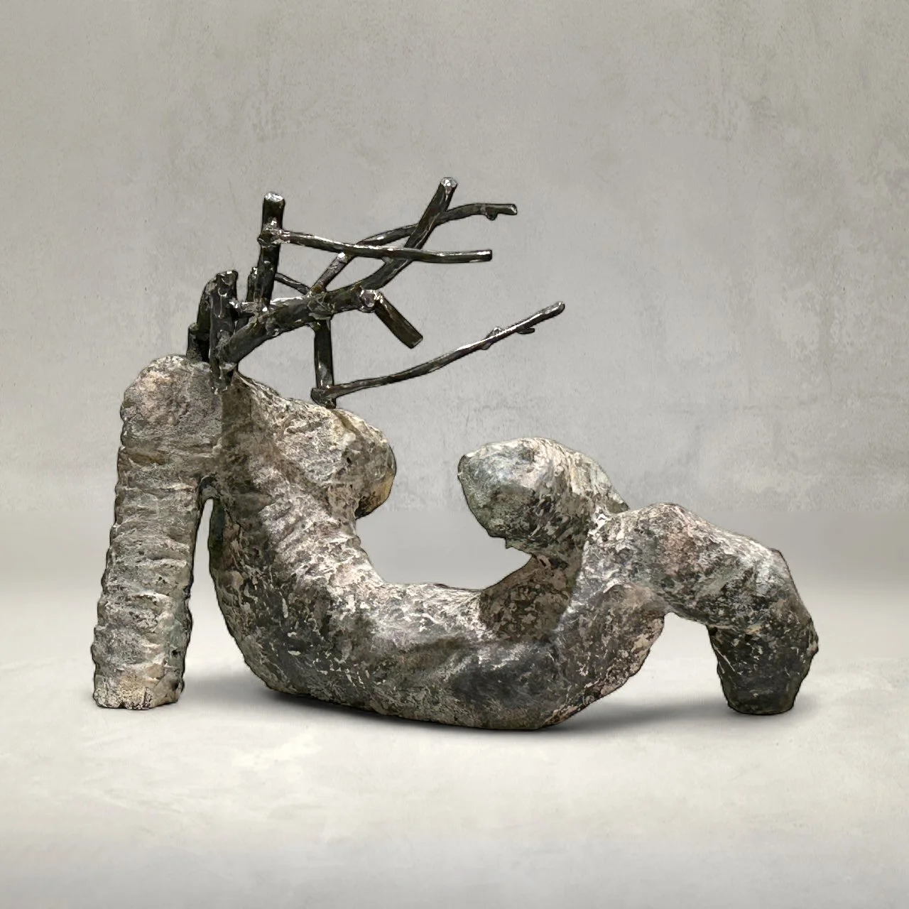 Sculpture of a curled-up figure with metal branches emerging from its back, mounted on a textured stone base.