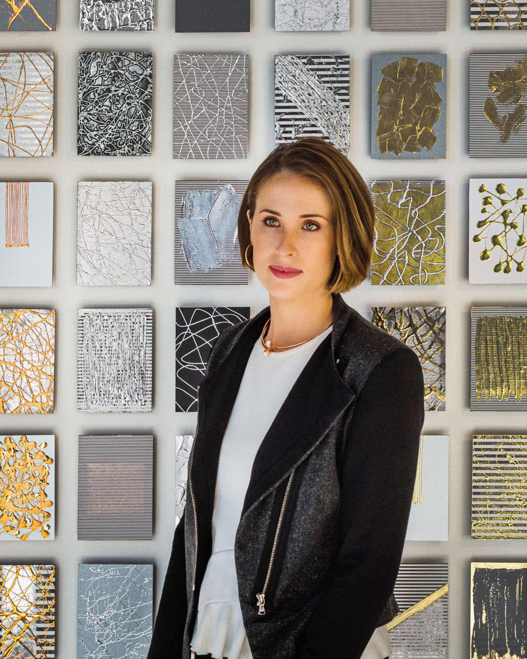 A woman with short brown hair and hoop earrings standing in front of a wall decorated with small framed abstract art pieces in silver, gold, black, and white colors.