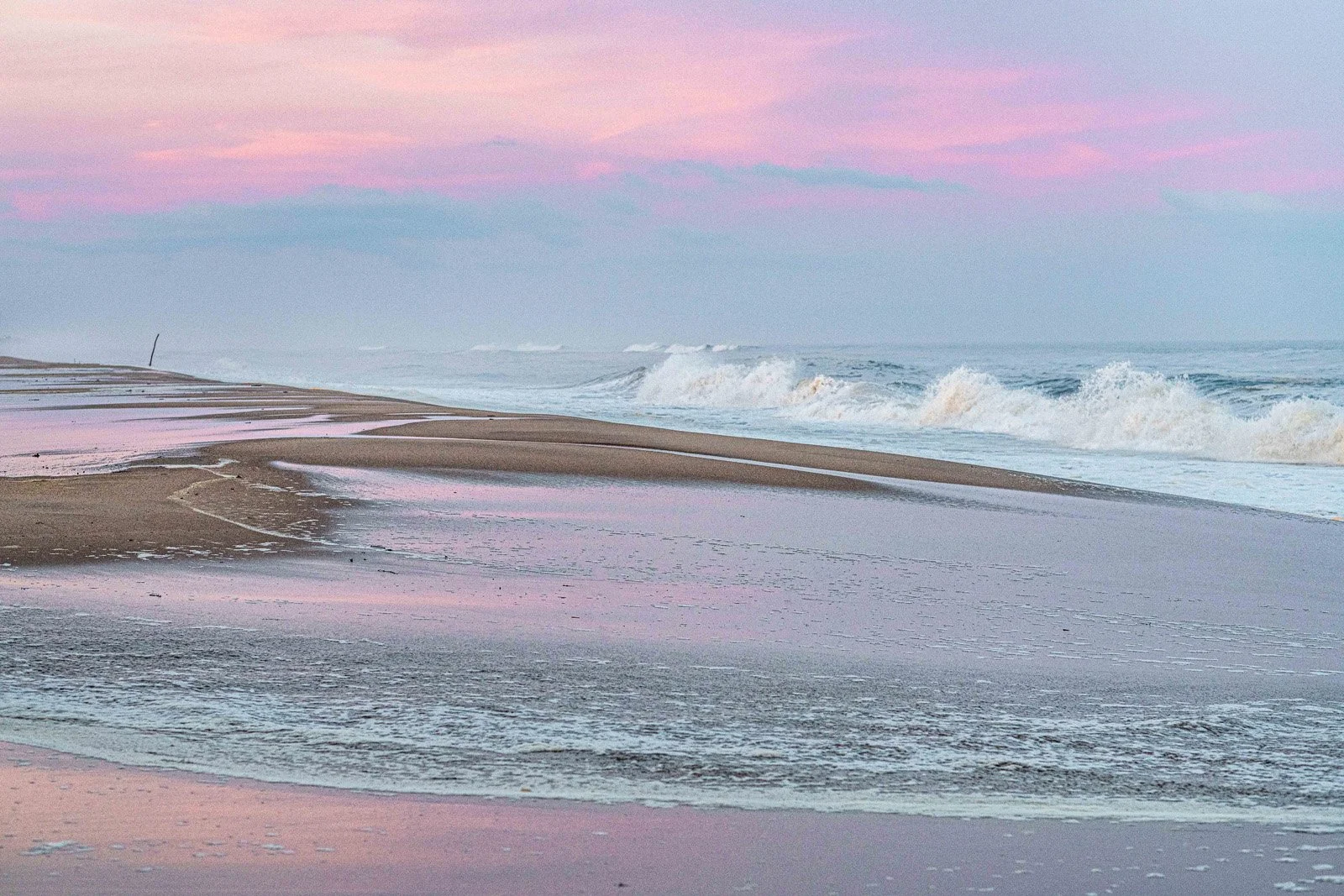 Empty beach with pink and purple sky, gentle waves crashing on the shore, wet sand reflecting the sky's colors.