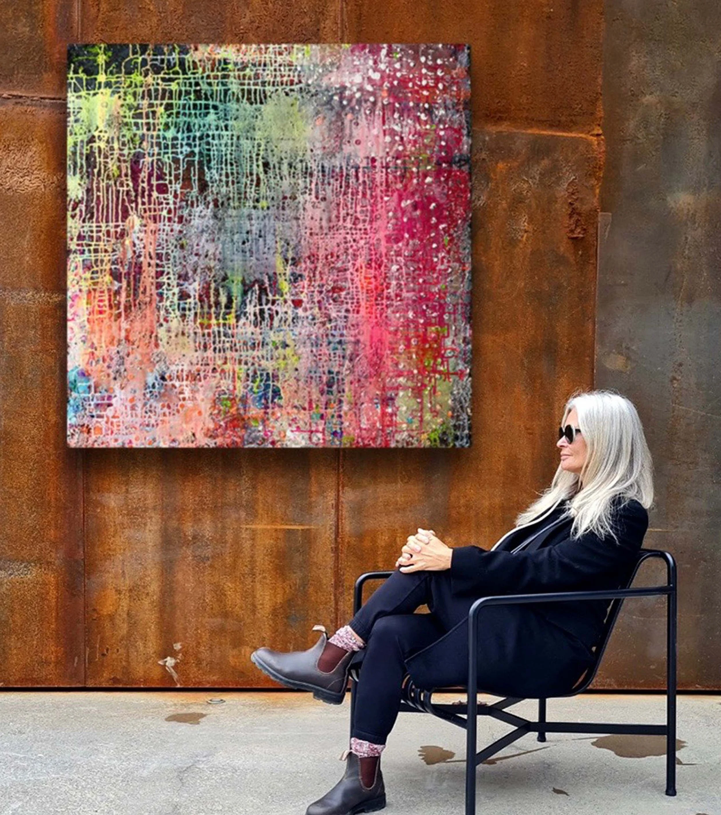 A woman with gray hair, sunglasses, and a black outfit is sitting on a black chair outside. She is positioned in profile, with her hands clasped and resting on her lap. Behind her is a large, colorful, abstract painting on a rust-colored wall.