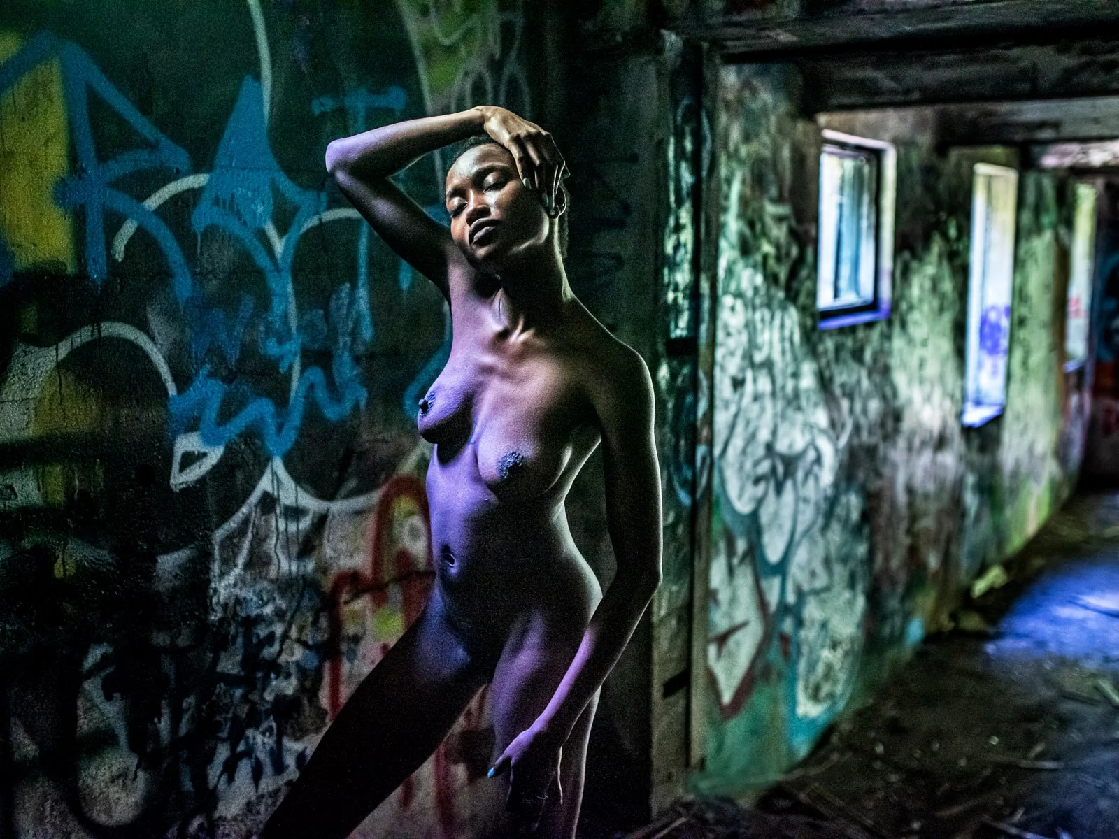 A nude woman with dark skin and short hair posing in a graffiti-covered urban setting with graffiti on the wall, standing with one arm raised and her hand on her head, with natural light coming from windows.