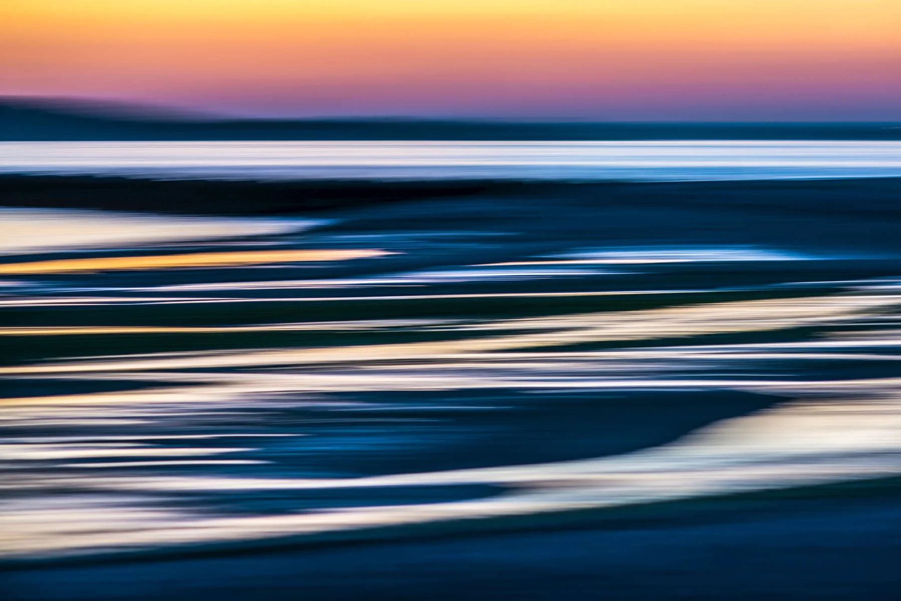 Abstract photo of a beach at sunset with blurred motion, showing the ocean waves and colorful sky with shades of pink, purple, and orange.