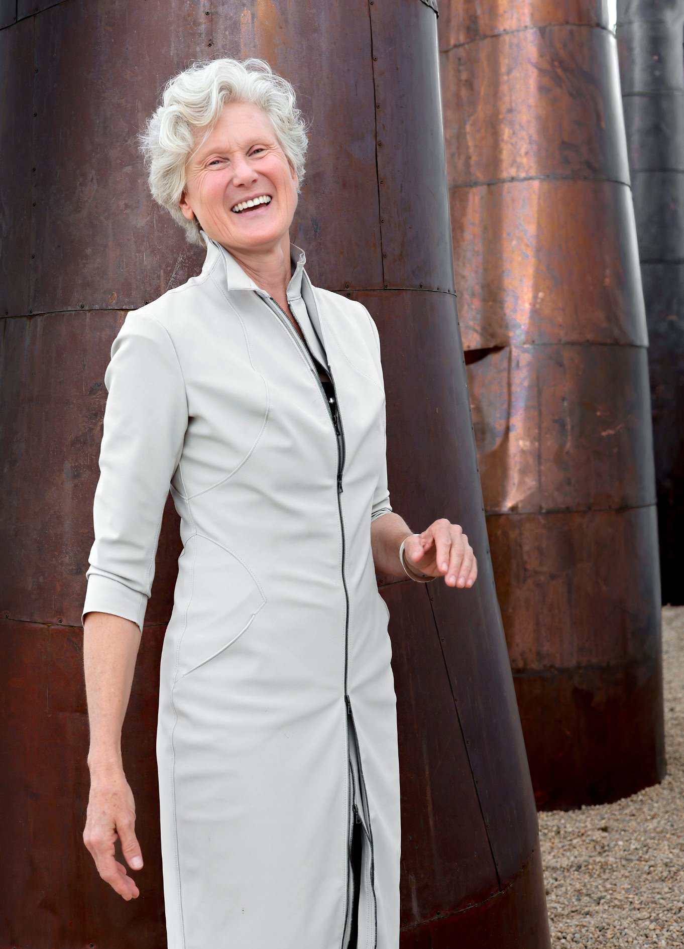 An older woman with short, curly gray hair smiling and standing outdoors in front of large, weathered, rusty metal structures.