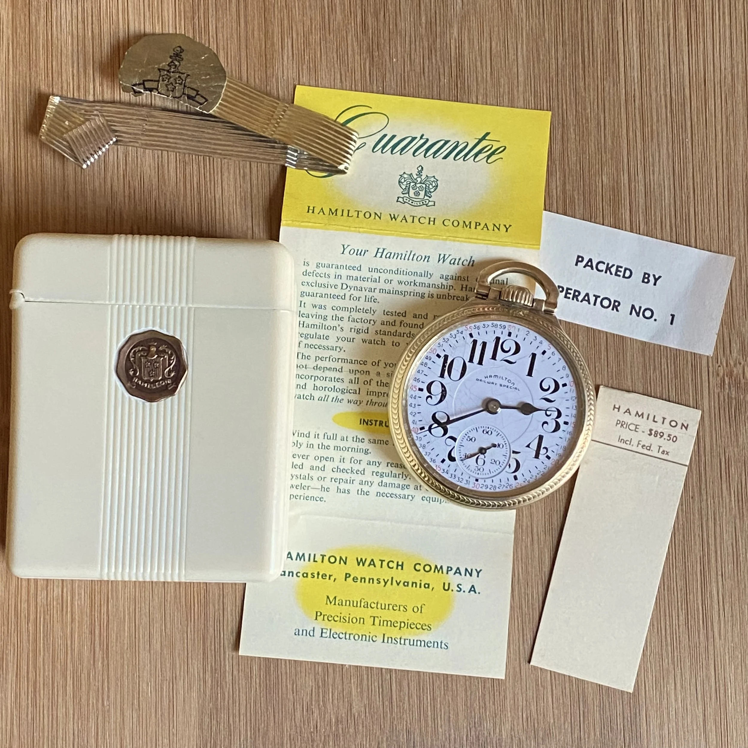Close-up image of a Hamilton watch with a white dial, gold case, and black numerals, placed on a wooden surface next to a yellow warranty card and some papers, with a ribbon cable and a white box visible partially.