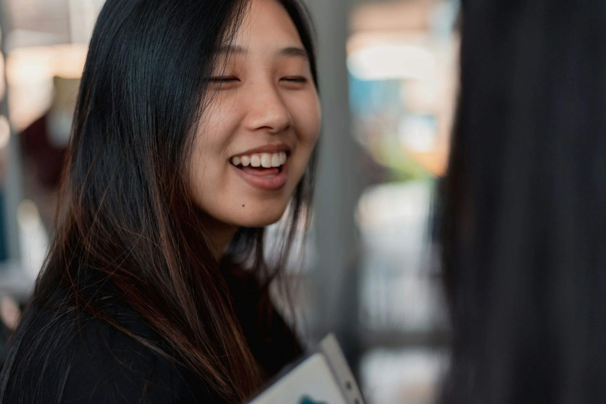 Close-up of a smiling young woman with long, dark hair, showing her teeth, in an indoor setting.
