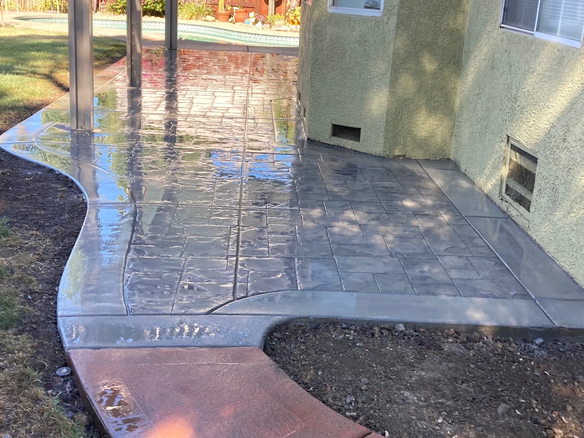 A wet concrete sidewalk with a curved design, adjacent to a yellow stucco house, reflecting nearby objects and sky, with an enclosed porch or patio area supported by metal posts.
