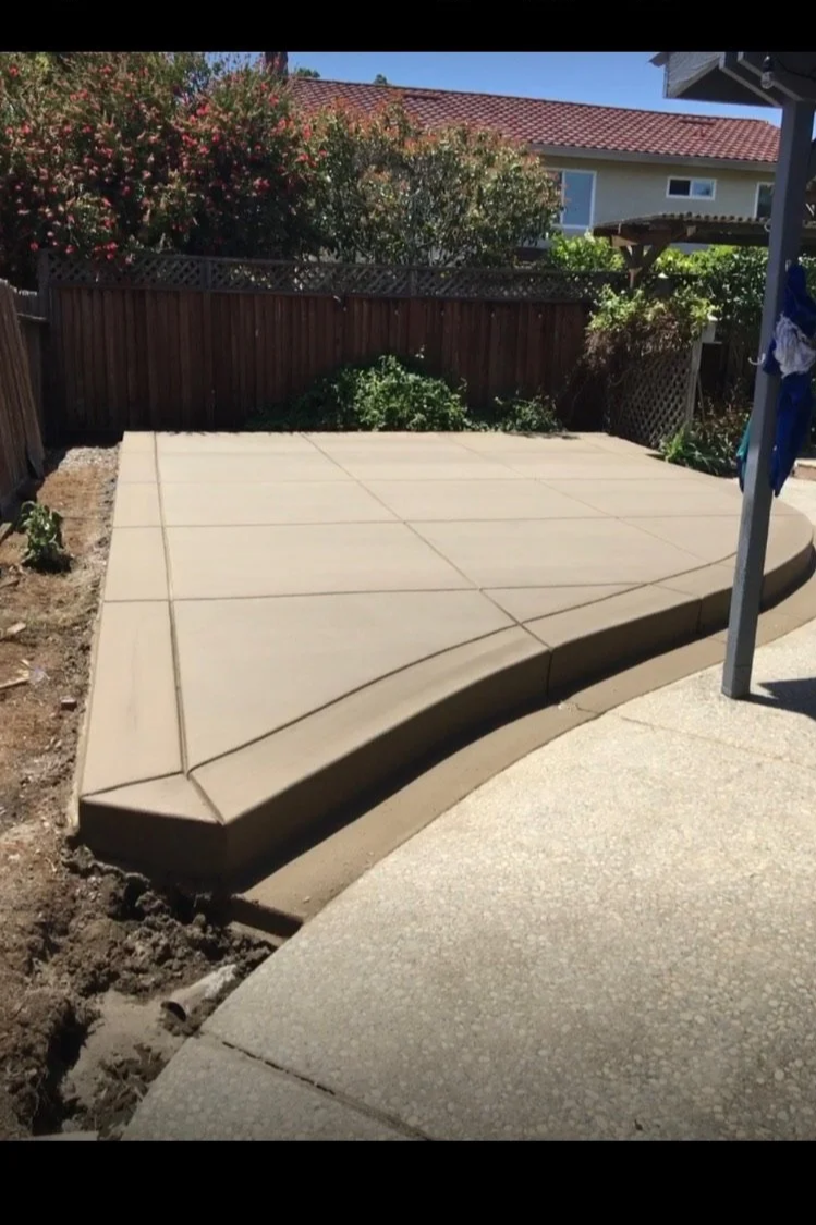 Image of a newly poured concrete patio with a decorative curved edge, surrounded by a garden with flowers and a wooden fence in the background.