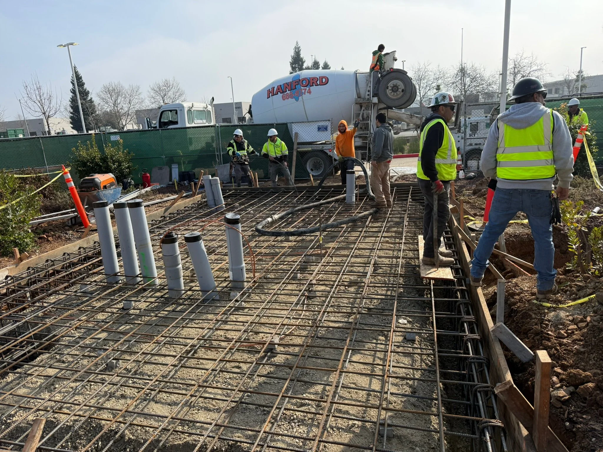 Construction workers at a site with rebar and pipes, some wearing safety vests and helmets, some standing near a concrete mixer truck labeled 'HANFORD' in red and blue, during daytime.