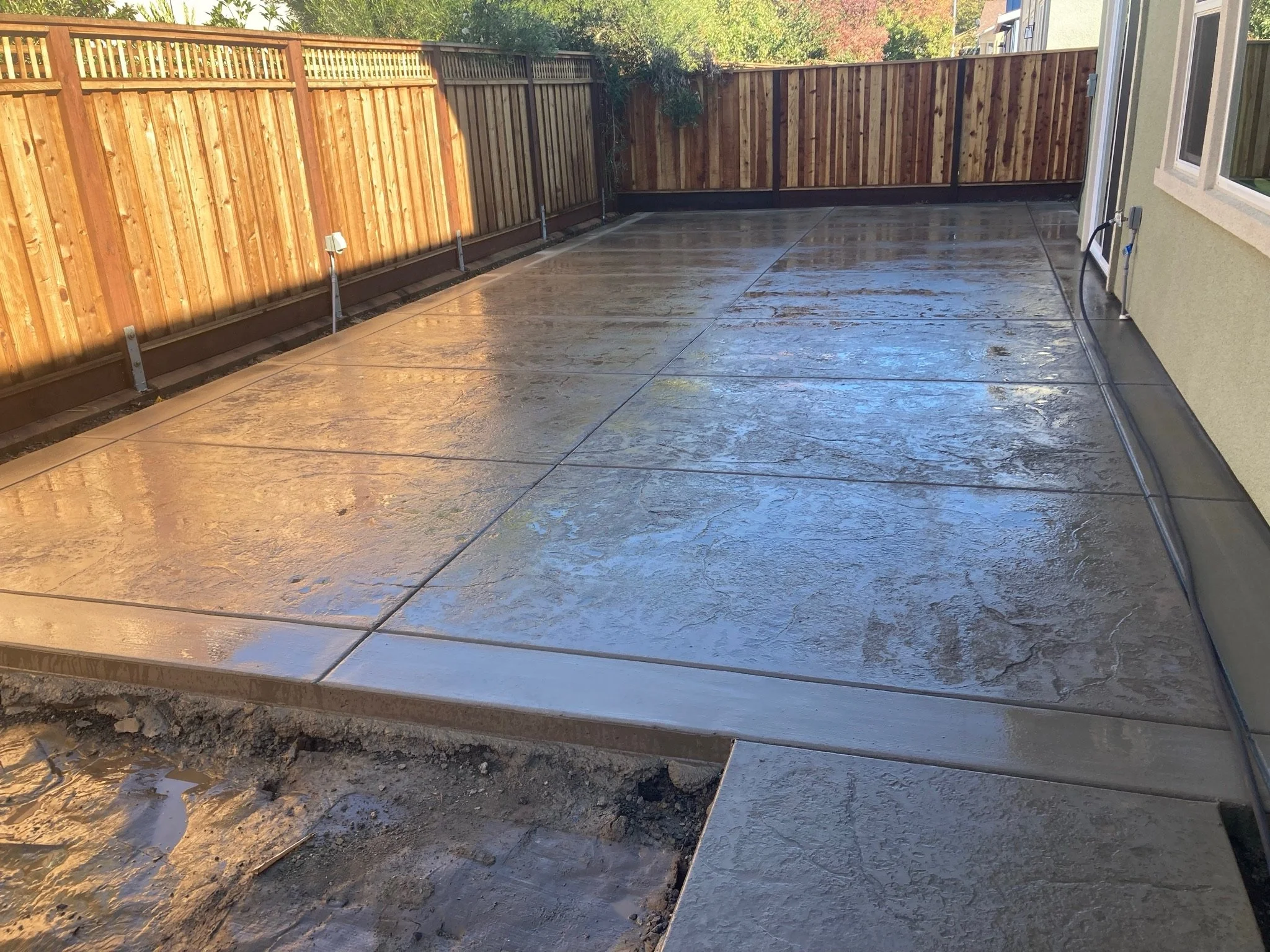 A newly installed concrete patio with some sections still wet, surrounded by a wooden privacy fence. The house exterior is visible on the right side.