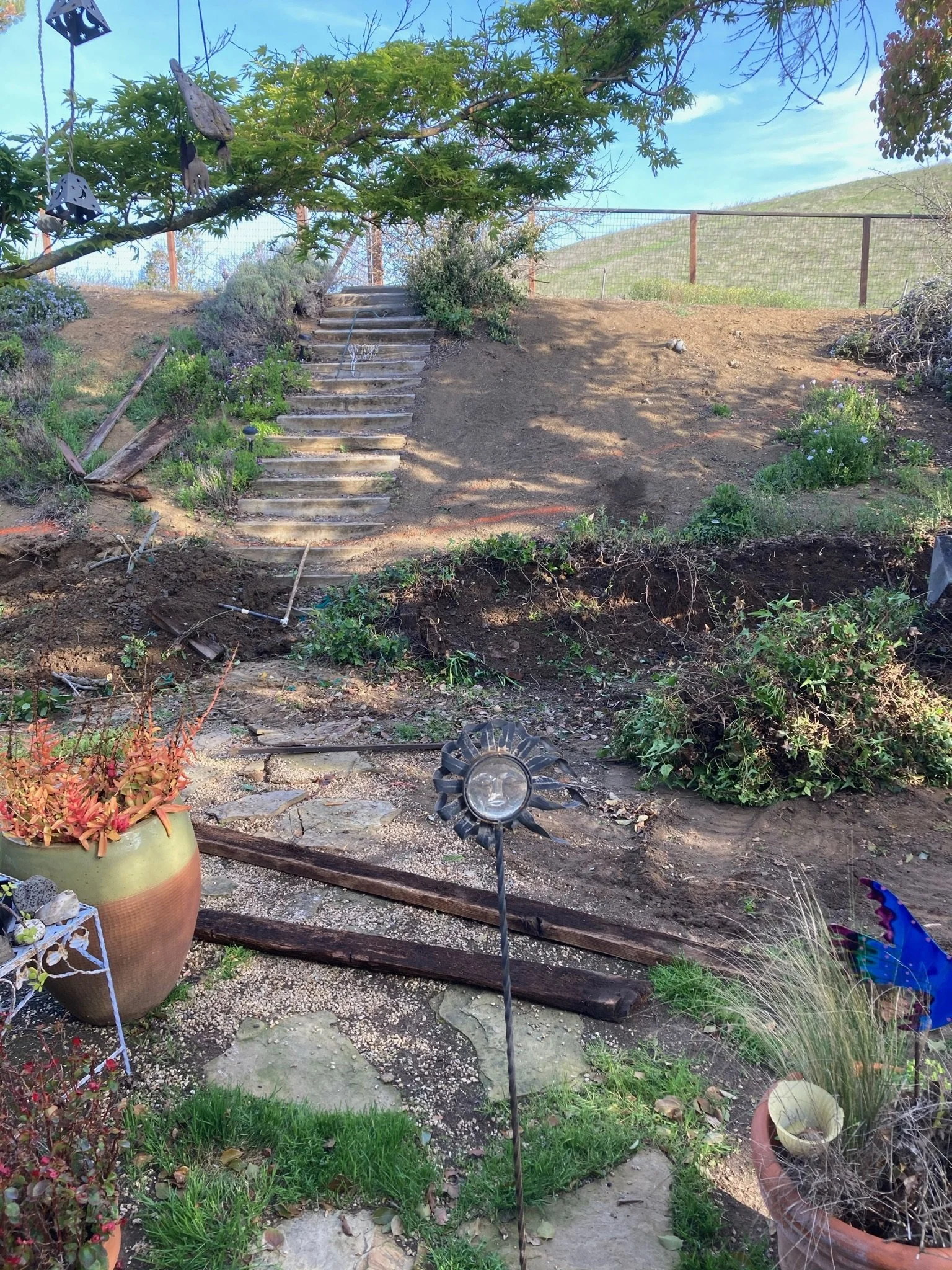A backyard garden scene with a metal sun sculpture, potted plants, a winding stone pathway, wooden steps, a tree with hanging decorations, and a wire fence on a hill in the background.