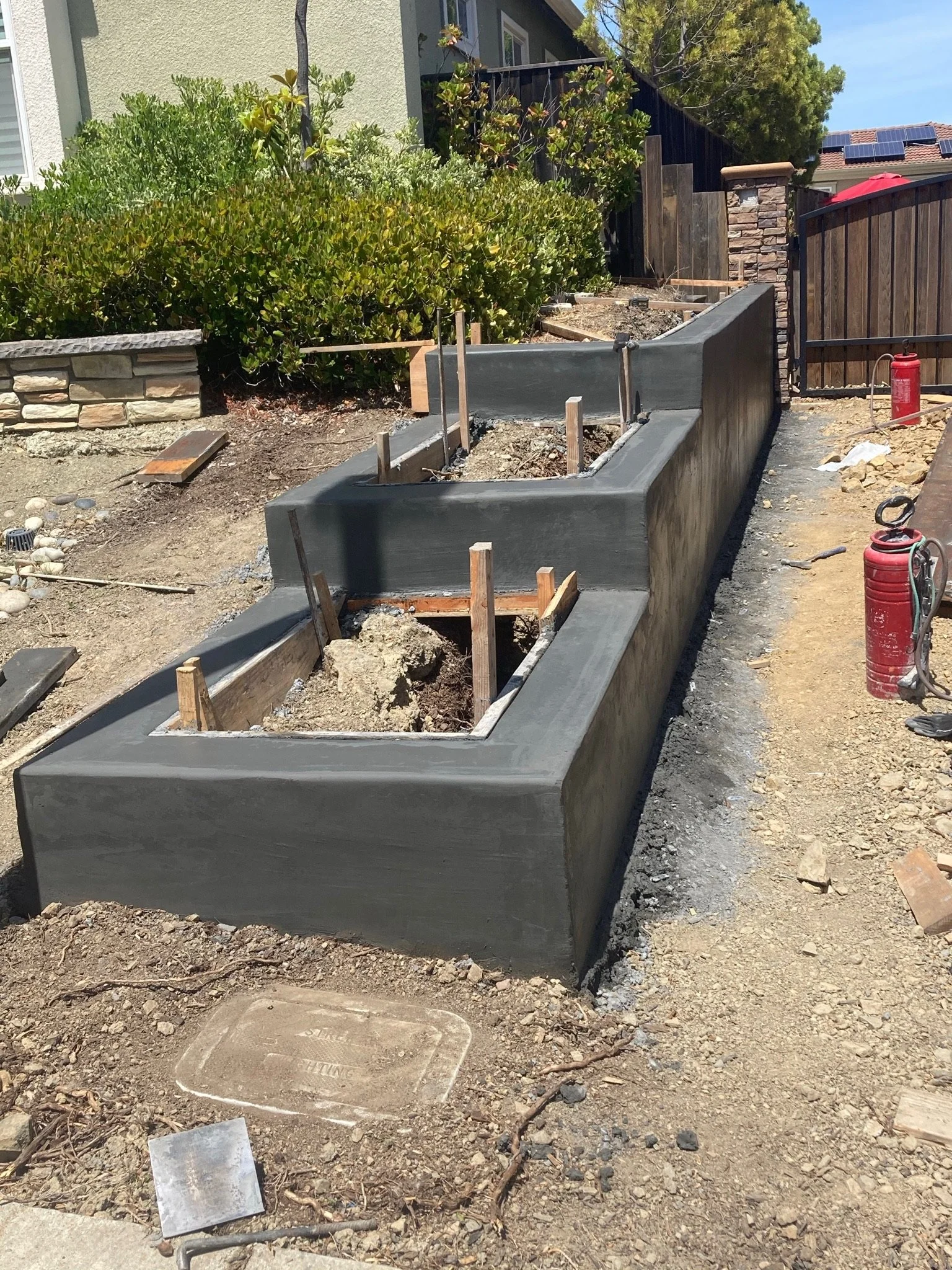 Under construction sidewalk with freshly poured concrete and wooden stakes, next to a stone wall and green bushes, in a residential backyard.