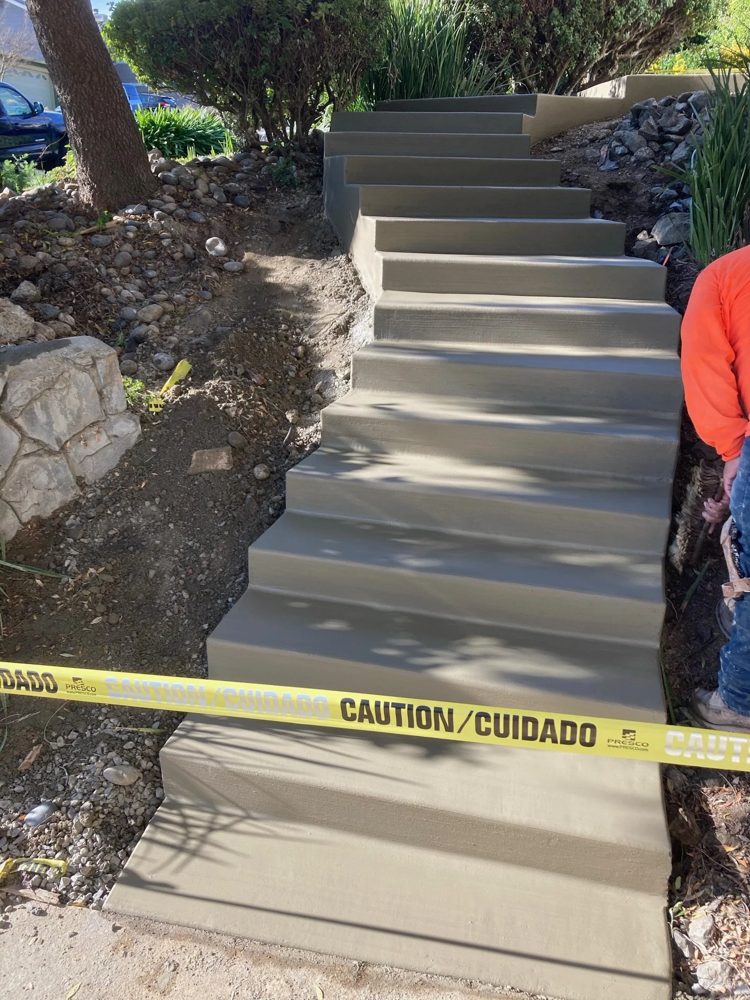 Concrete stairs under construction with caution tape in front, surrounded by dirt and rocks, with plants and bushes in background.