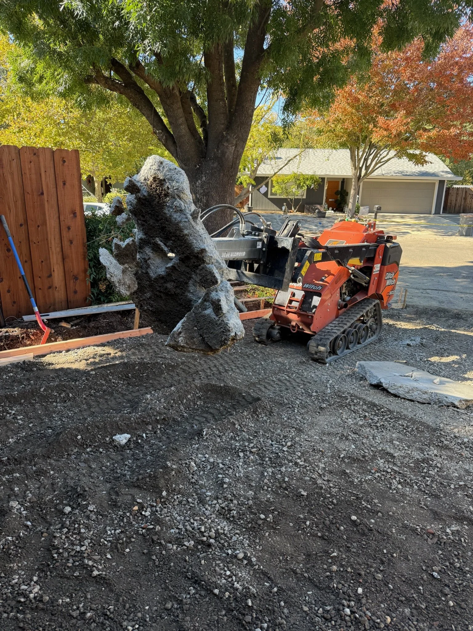 A backyard with a large tree and a wooden fence on the left, a construction vehicle lifting a large rock, and a house with a garage in the background, with autumn trees and a partly paved driveway.