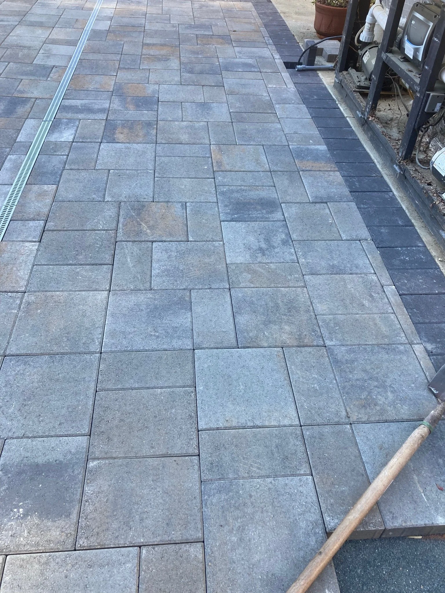 Recent installation of outdoor pavers in gray and black, with a drainage grate along the edge, and construction tools nearby.