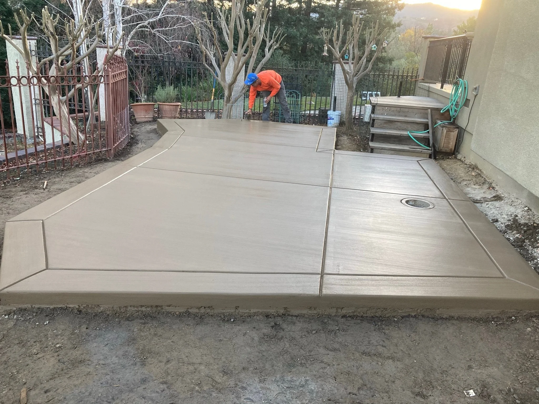 A person in an orange jacket and blue cap working on installing a large concrete patio in a backyard, surrounded by trees, a fence, and some potted plants, with steps leading to a house with a garden hose coiled on the side.