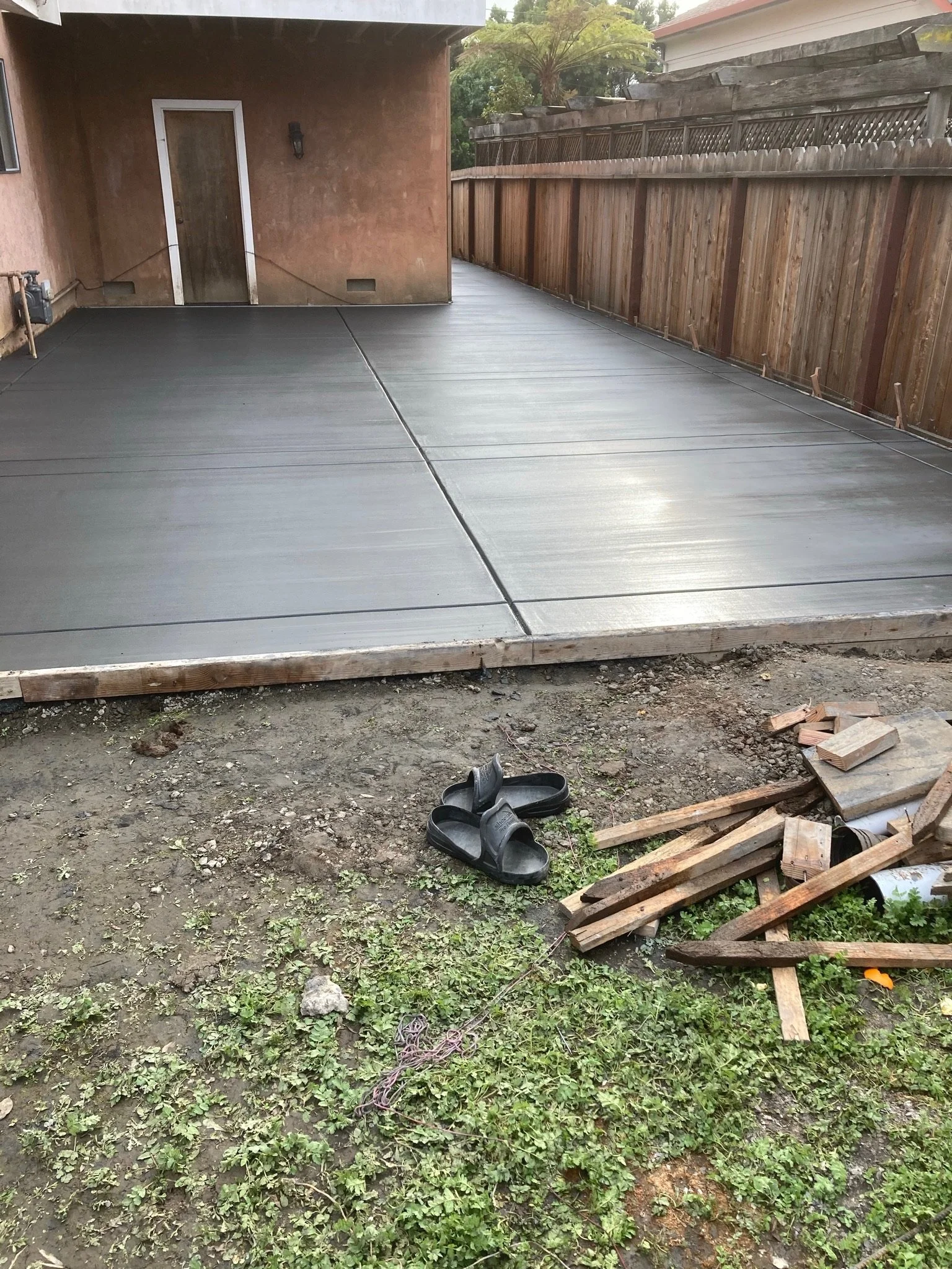 A small backyard patio with a newly poured concrete slab, a black pair of slippers on the dirt, some scattered wood planks, a rope, and small plants growing in the soil.