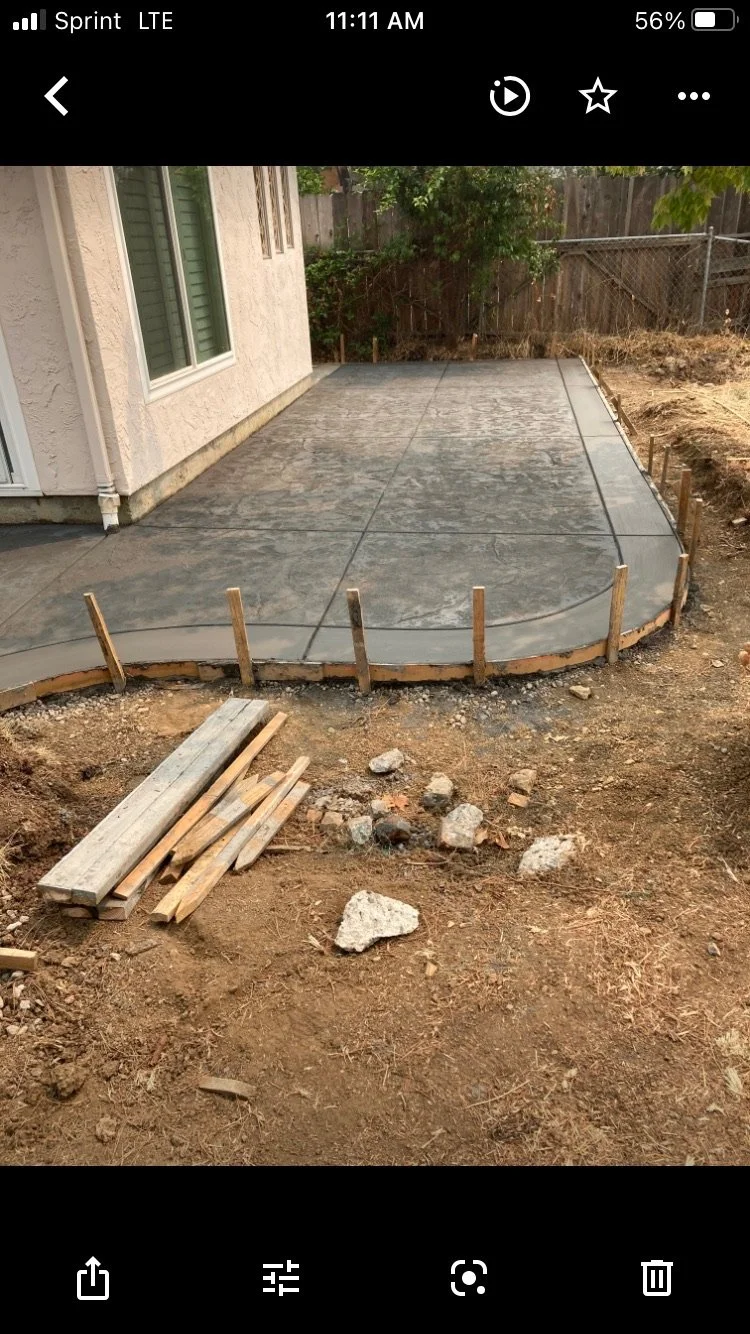 Concrete patio being constructed outside a house with partially completed curved edges, surrounding dirt and construction supplies.