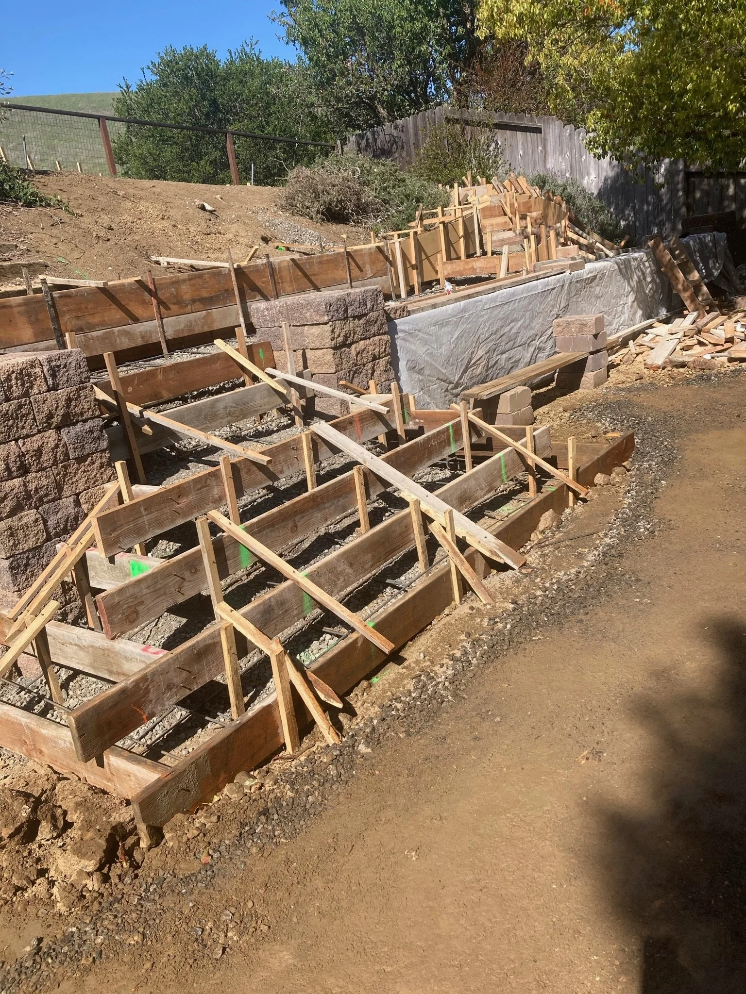 Construction site with wooden forms and retainers for concrete, along a dirt pathway and a stone wall, under a blue sky with trees.