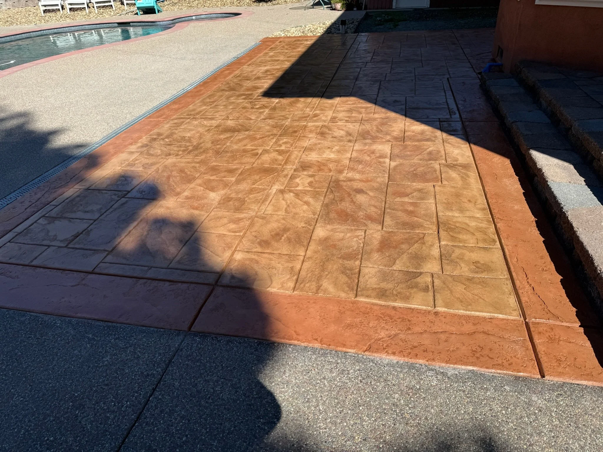 Outdoor scene showing a swimming pool, patio with different types of paving, and some patio furniture in the background.