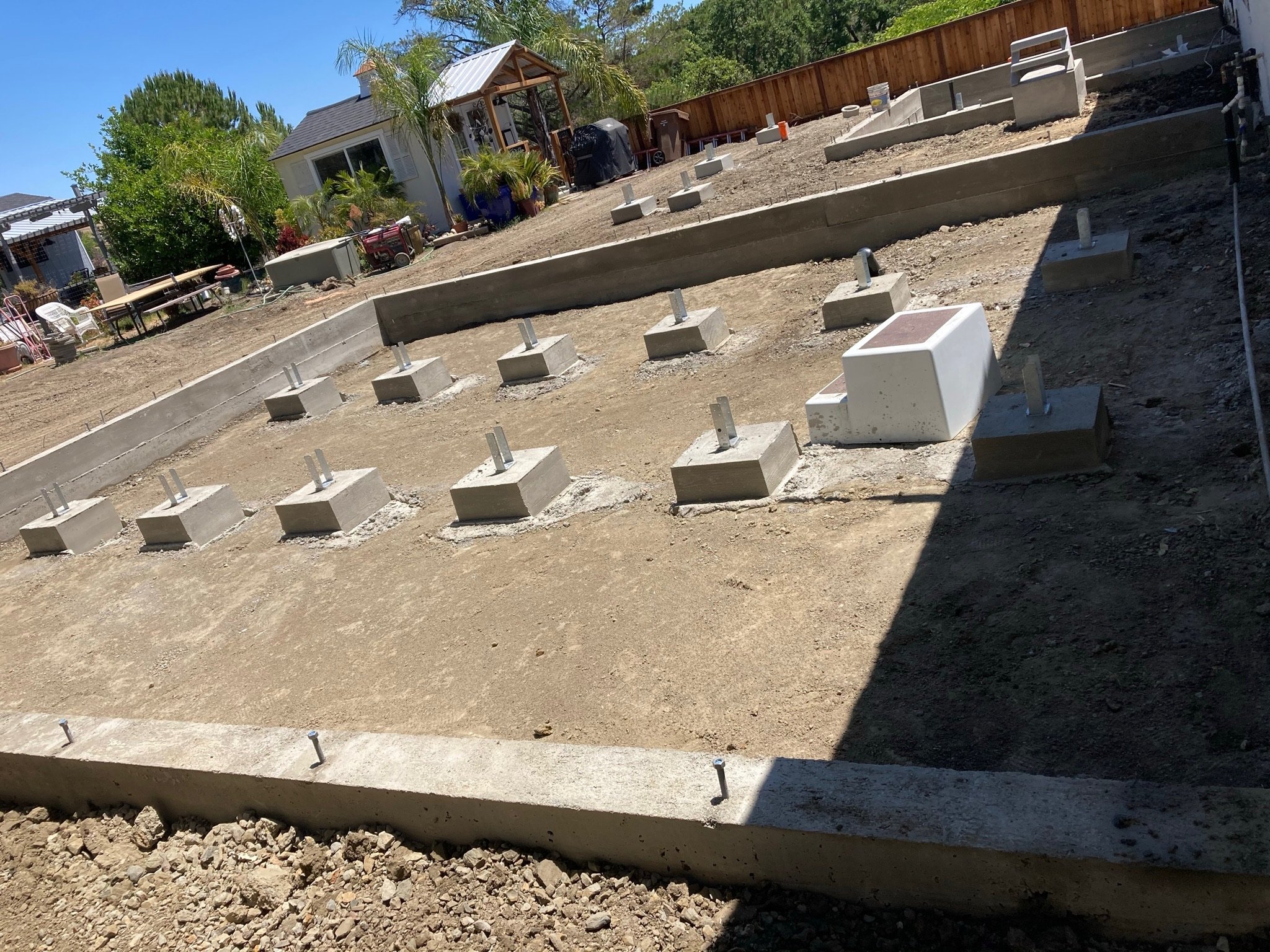 Backyard with construction in progress, concrete patio foundation with metal fasteners, dirt ground, plants and trees, house in the background.