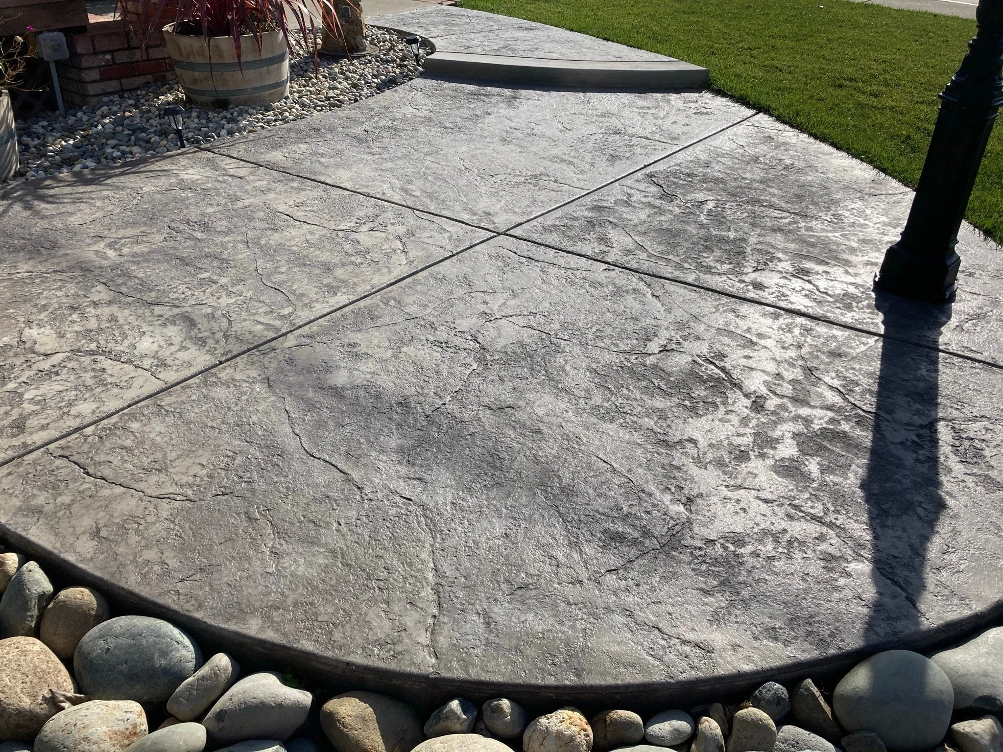 Concrete sidewalk with a step leading to grass area, surrounded by decorative rocks and pebbles, with some plants and garden lights nearby.
