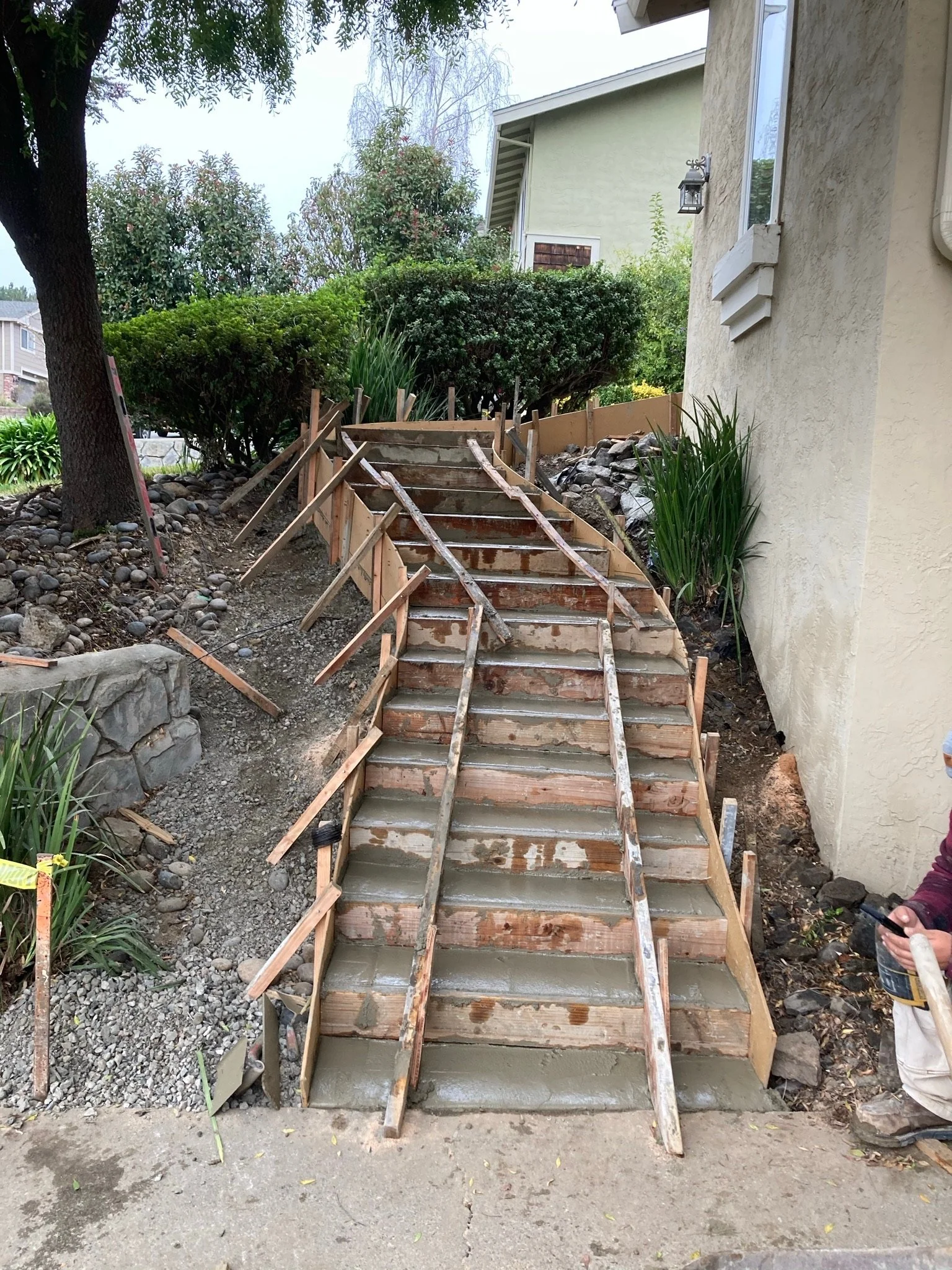 A wooden staircase under construction with concrete being poured into the steps, next to a beige house wall and surrounded by greenery. There is a person wearing white pants and a gray jacket holding a phone on the right side.