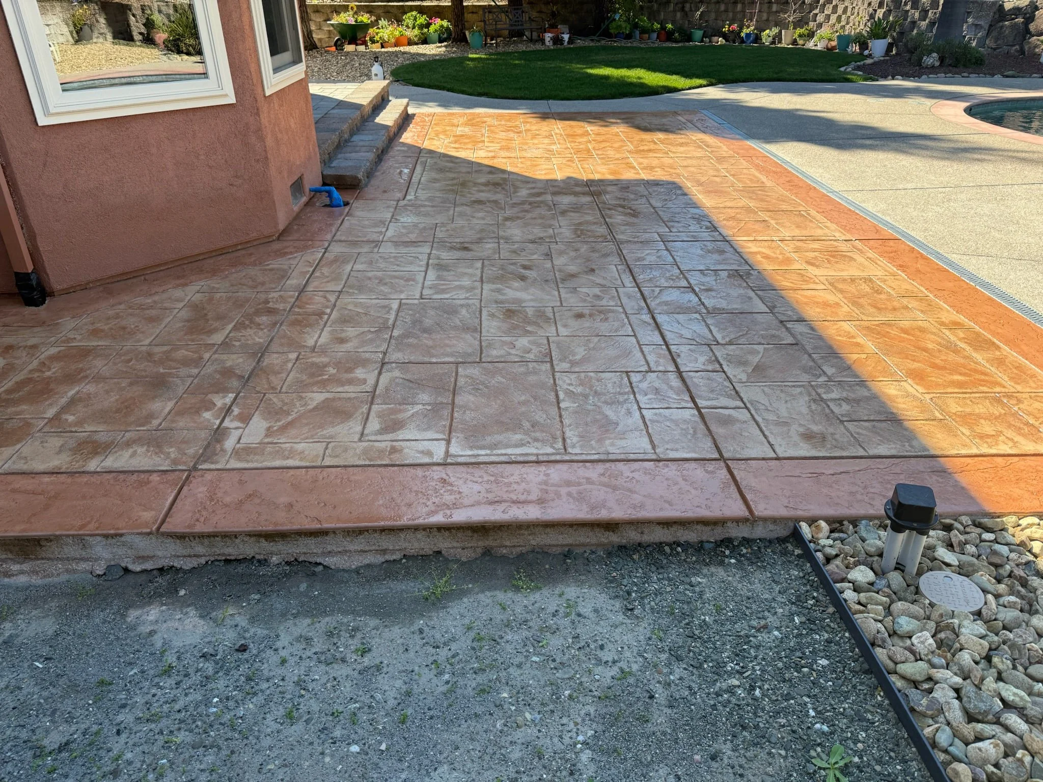 Backyard patio with newly installed stamped concrete in shades of brown and tan. The patio has steps leading up to a grassy area with potted plants along a stone wall. There are some garden tools and a small section of exposed soil in the foreground.