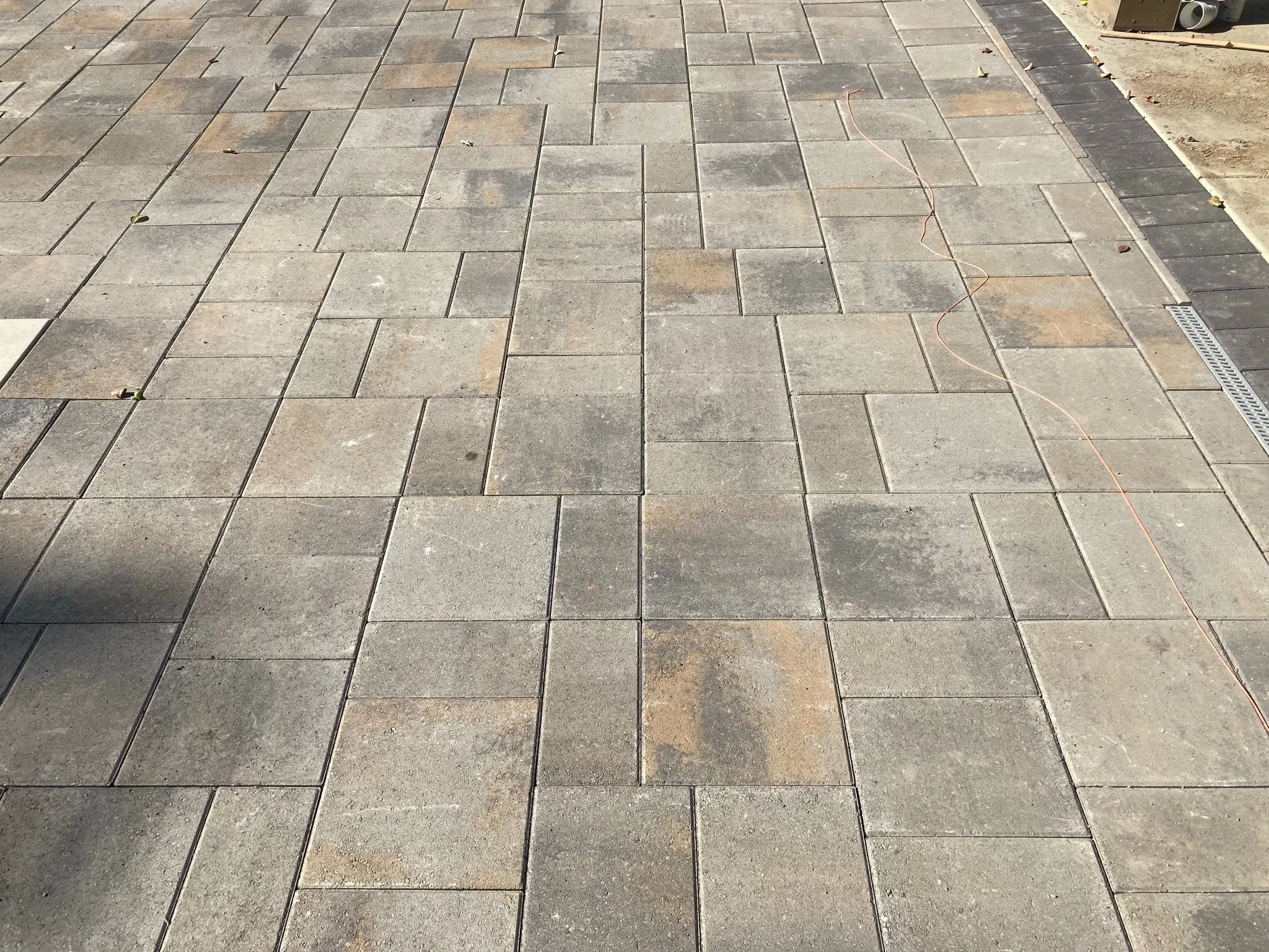 Close-up of a paved walk with rectangular gray stones arranged in a pattern, with some leaves and a thin orange wire or string running across the pavement, and a drainage grate visible on the right edge.