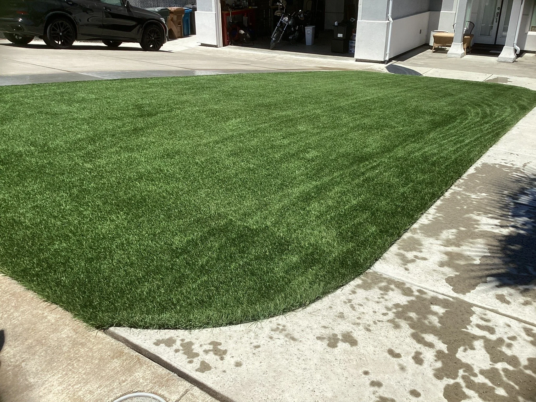 A freshly laid green sod lawn with a curb shadow in the foreground and a concrete sidewalk at the edge. A driveway with a black car and garage in the background, with visible storage items and a motorcycle.