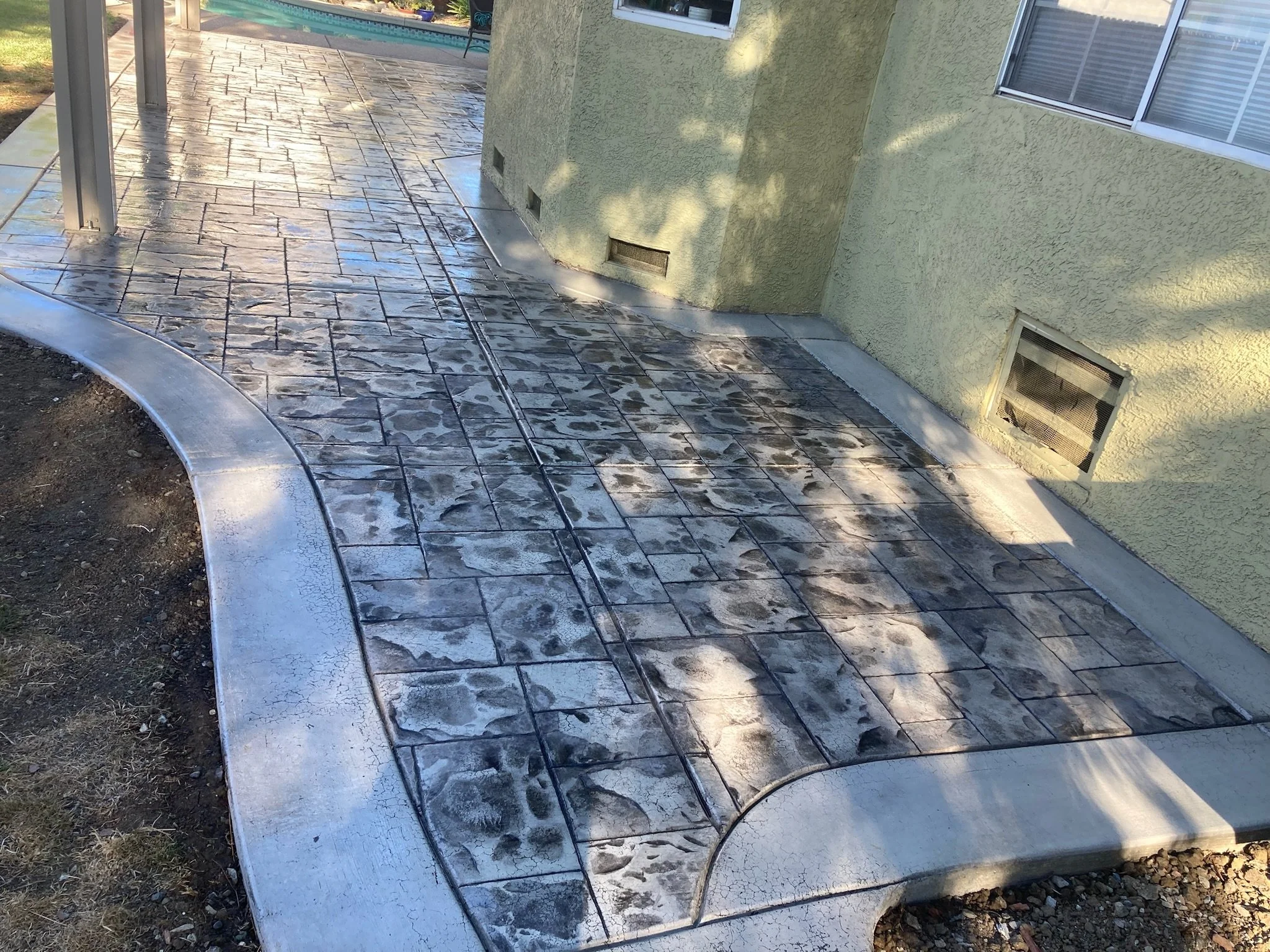 A backyard patio with stamped concrete flooring that looks like irregular stone tiles, wet from recent cleaning, beside a yellow stucco house with vents near the ground and large sliding glass windows. A landscaped area with grass and a swimming pool