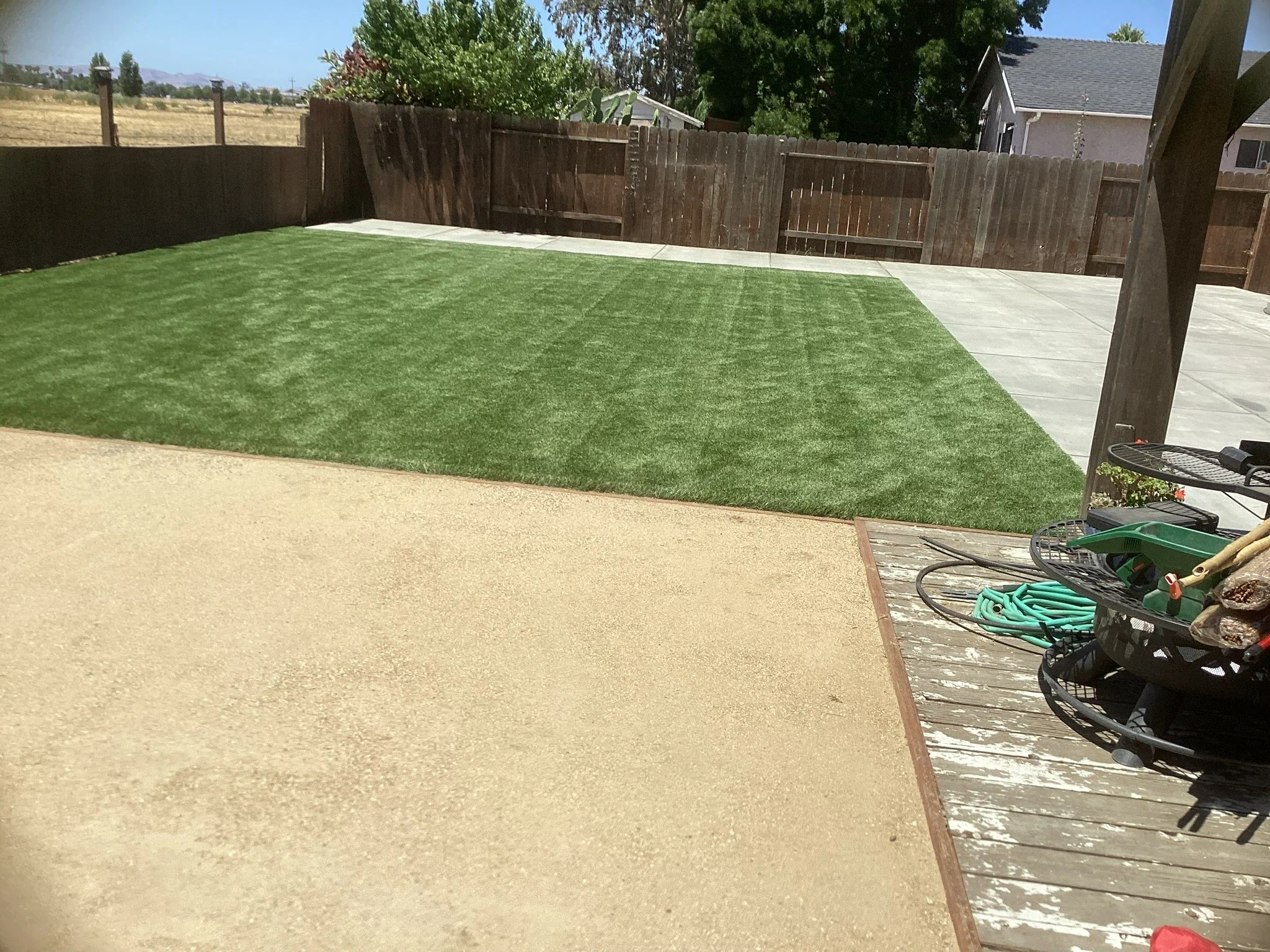 A backyard with a freshly laid artificial grass area, adjacent to a concrete patio, surrounded by a wooden fence; garden tools are visible on a small table in the corner.
