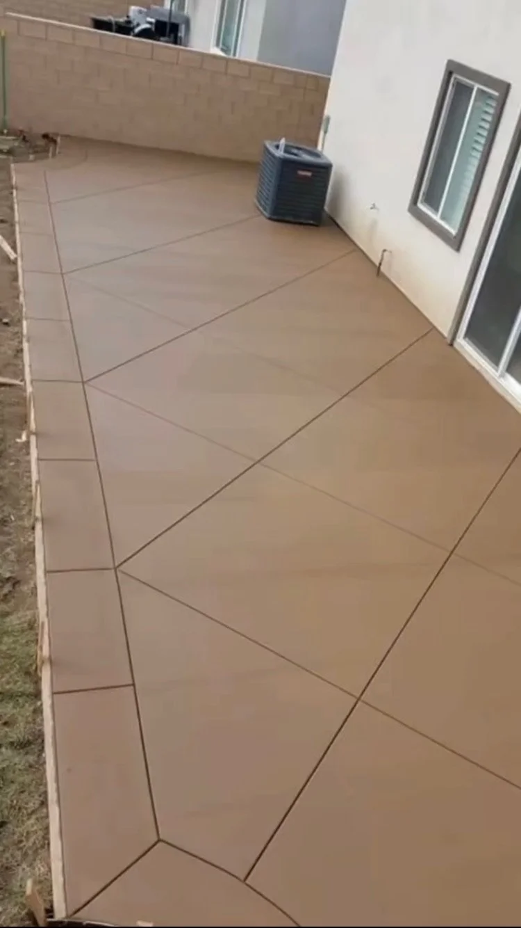 Newly installed backyard concrete patio with patterned scoring, beige color, next to a white house wall with a window, and an air conditioning unit, with a dirt area on the left side.