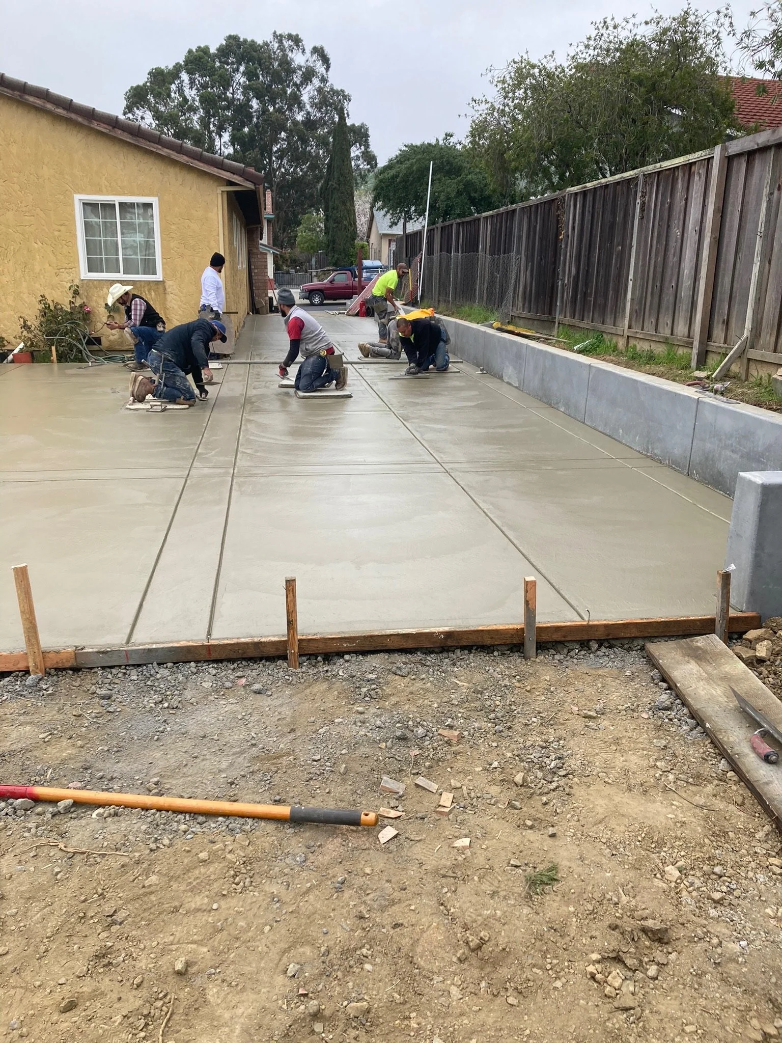 Construction workers pouring and smoothing concrete on a driveway next to a yellow house, with tools and a wheelbarrow visible.