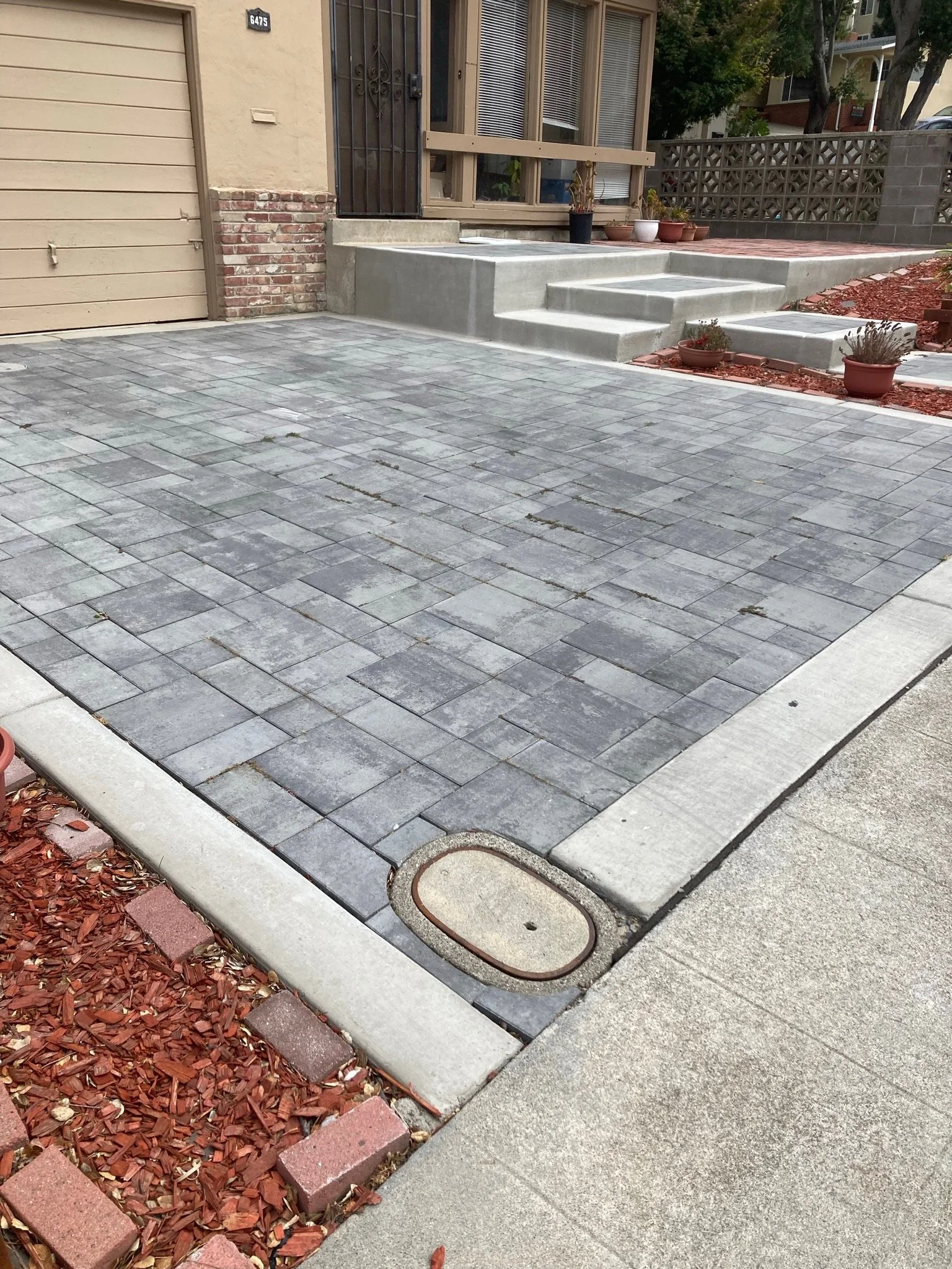 Front entrance with concrete steps, potted plants, a metal security door, and a beige garage door. The ground is paved with gray bricks, and there is a small garden bed with red mulch and bricks.
